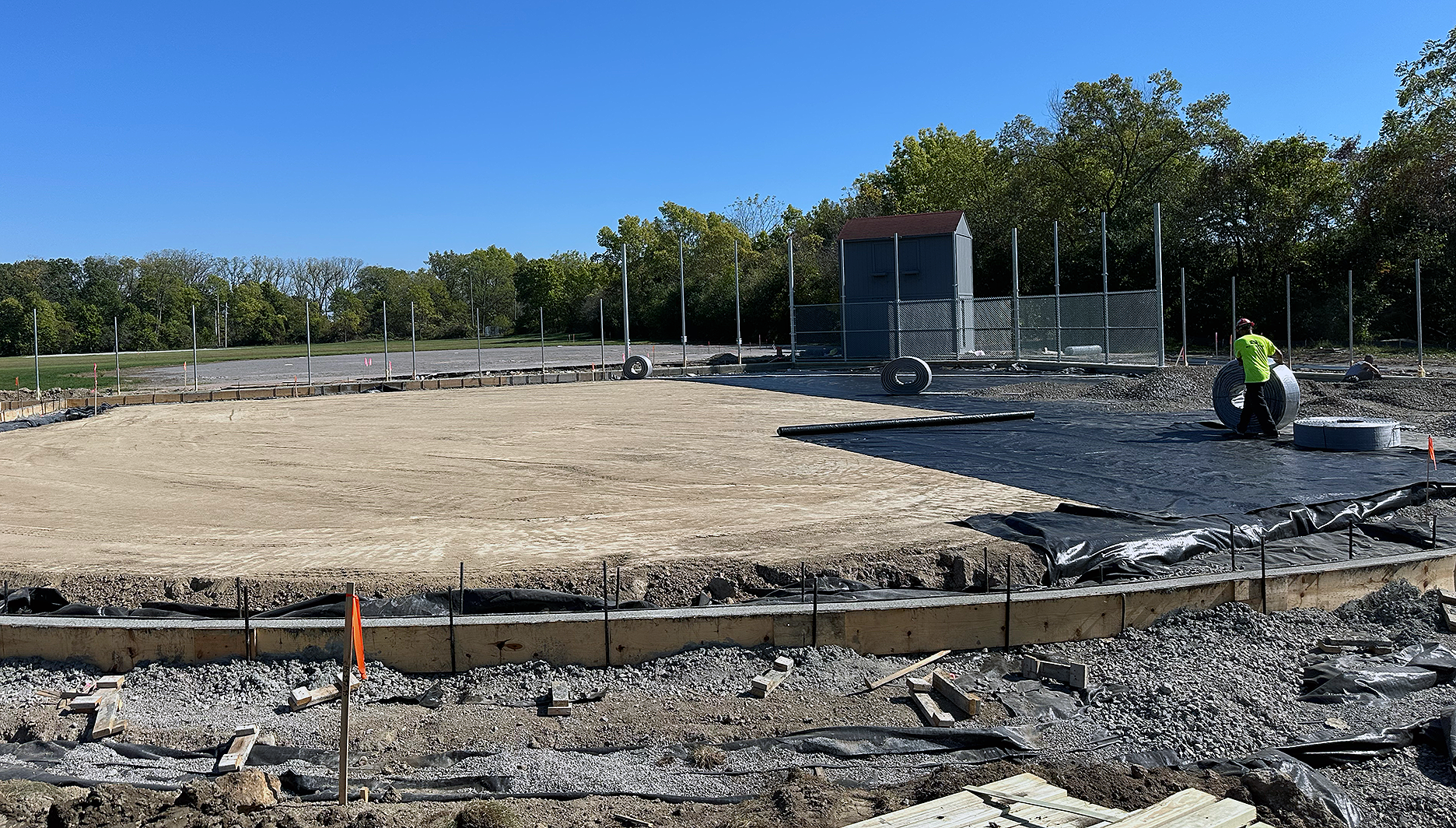 Construction site with a large, leveled dirt area, black plastic sheeting, and a worker laying a concrete surface, with trees and a blue sky in the background.