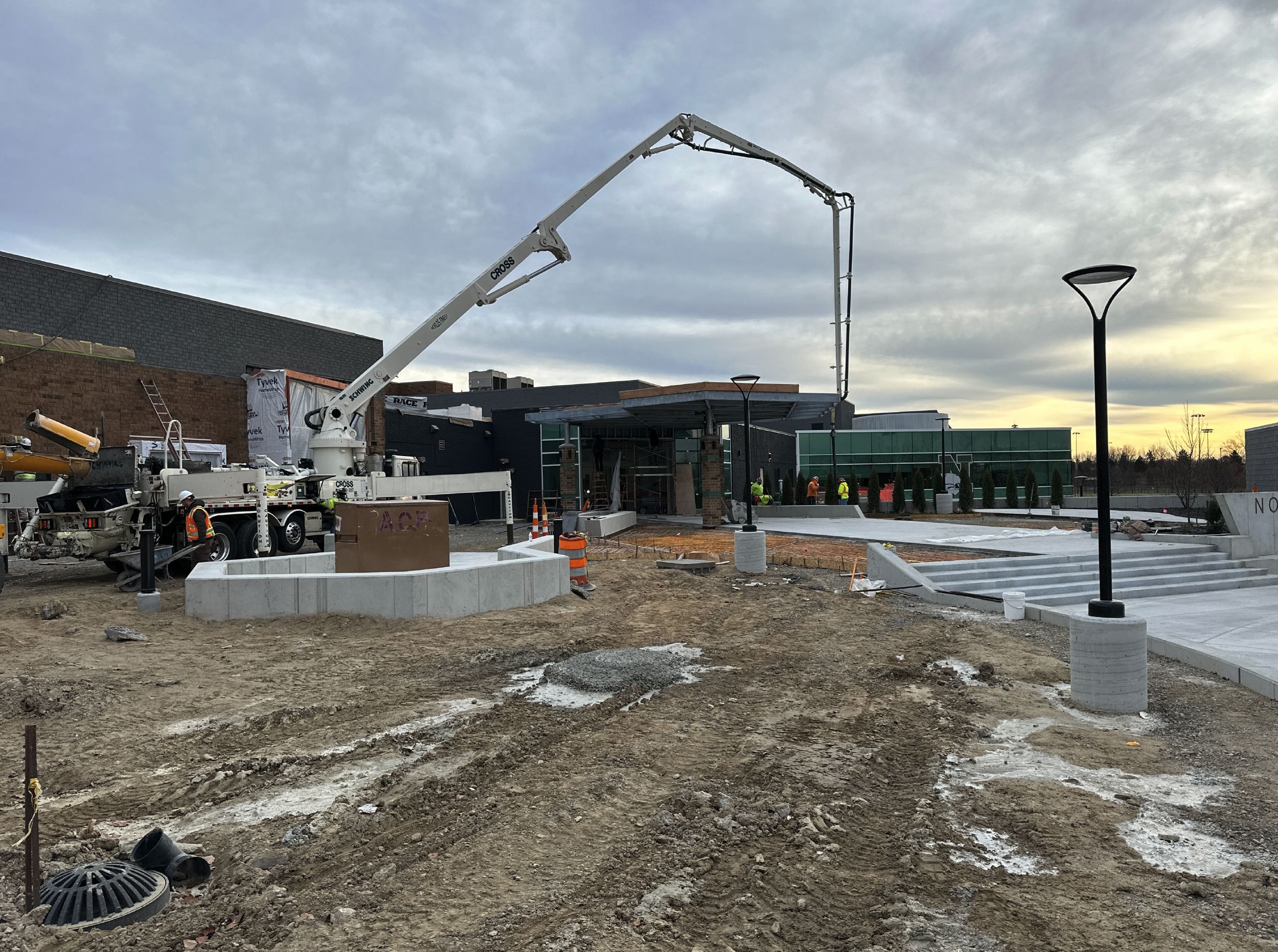 Construction site with workers, concrete structures, a crane, street lamps, and a building in the background, during sunset.