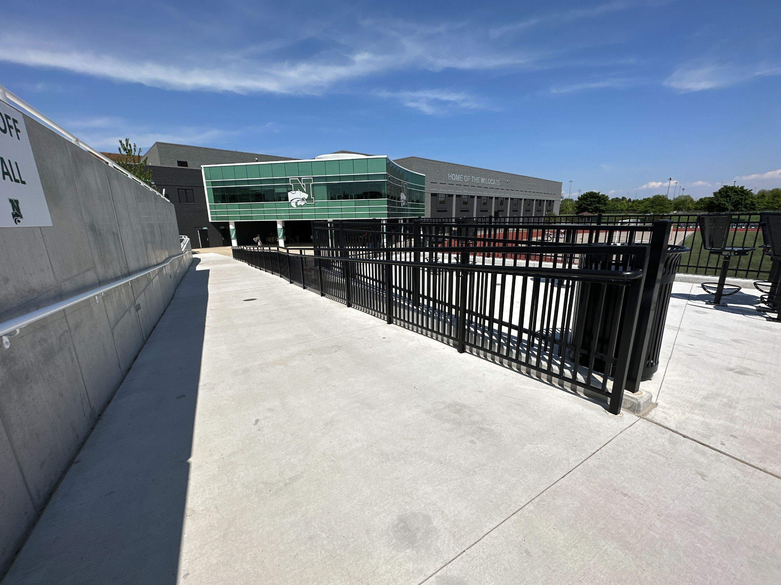 Exterior view of Novi High School building highlighting a high quality concrete construction project by Simone Companies with green and gray facade, sports field and bleachers, clear blue sky, and sidewalk with black metal fencing.