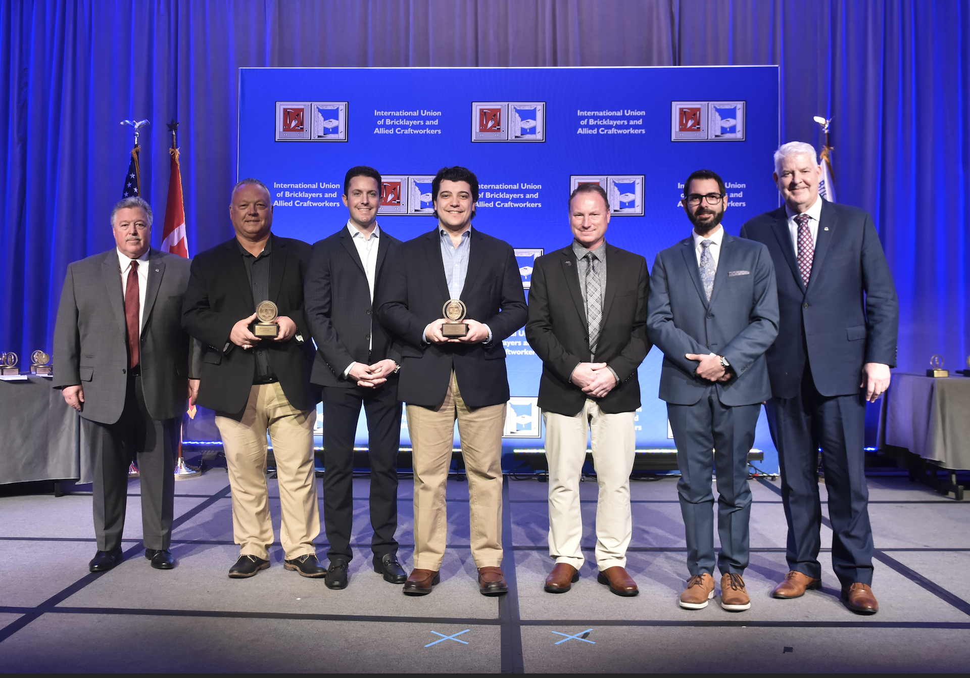 Group of seven men standing on stage at an award ceremony, three holding trophies, backdrop displaying logos and text for the International Union of Bricklayers and Allied Craftworkers.