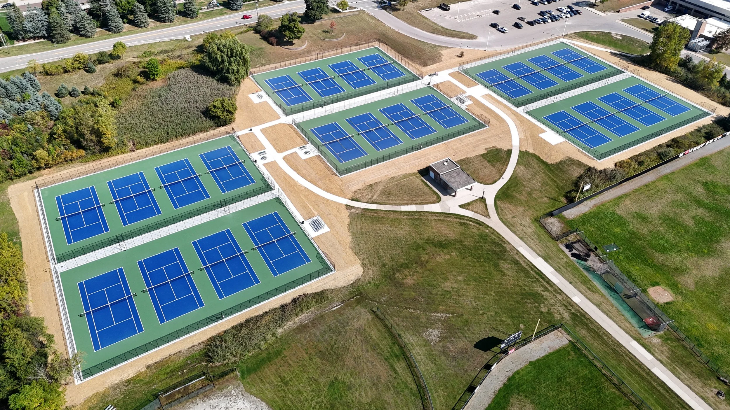 An aerial view of multiple tennis courts with blue surfaces and black fencing, surrounded by green fields, trees, and parking lots.