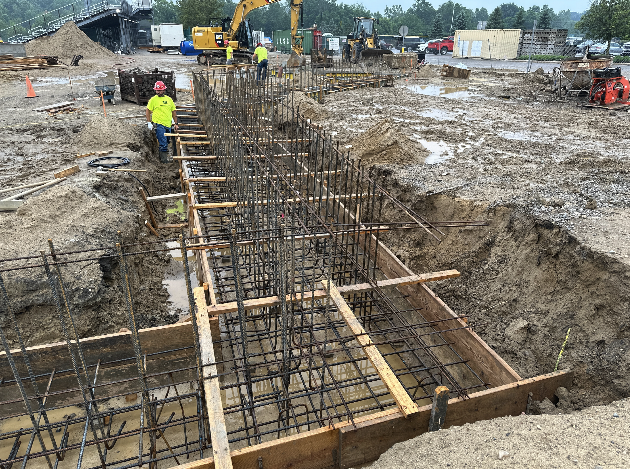 Construction workers in bright yellow shirts and helmets working on rebar framework inside a large trench at a construction site, with heavy machinery, dirt piles, and parked cars in the background.