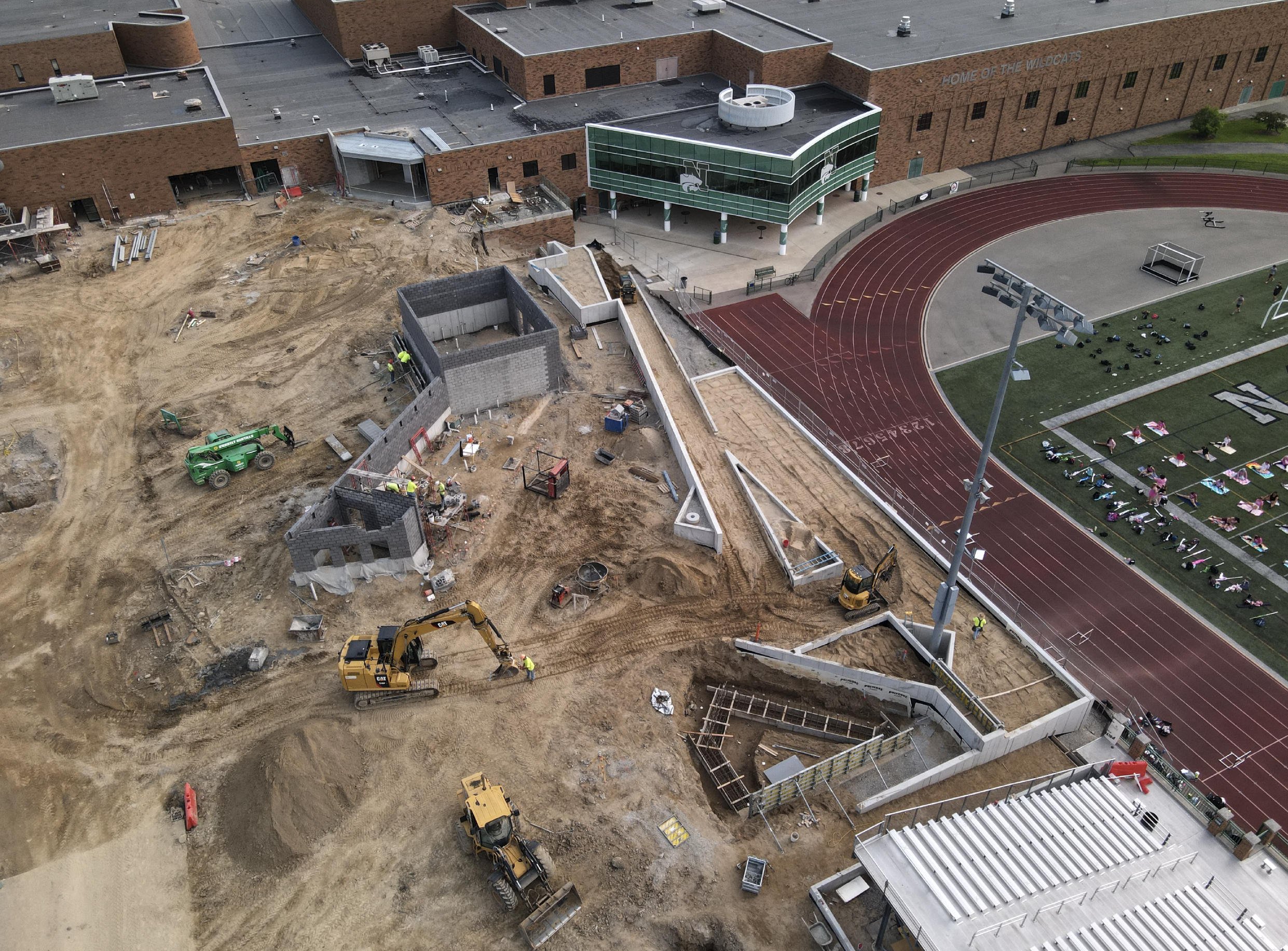 Construction site next to a school's running track and football field, with workers operating machinery and constructing walls and structures.