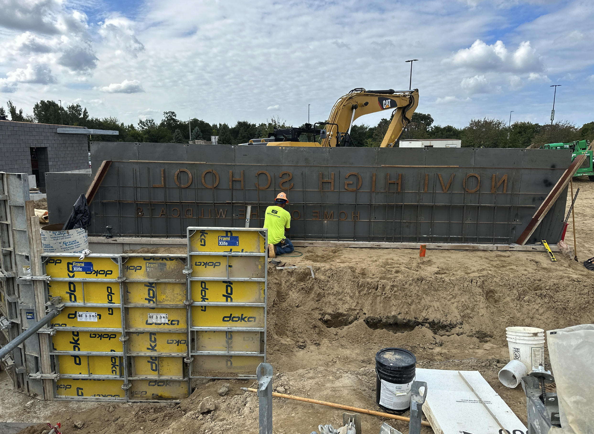 Construction site with a worker wearing a neon yellow shirt and orange safety helmet, working on a large steel formwork. There is a yellow Caterpillar excavator in the background, and steel reinforcement bars are visible. The area is partly excavated with dirt and construction materials scattered around.