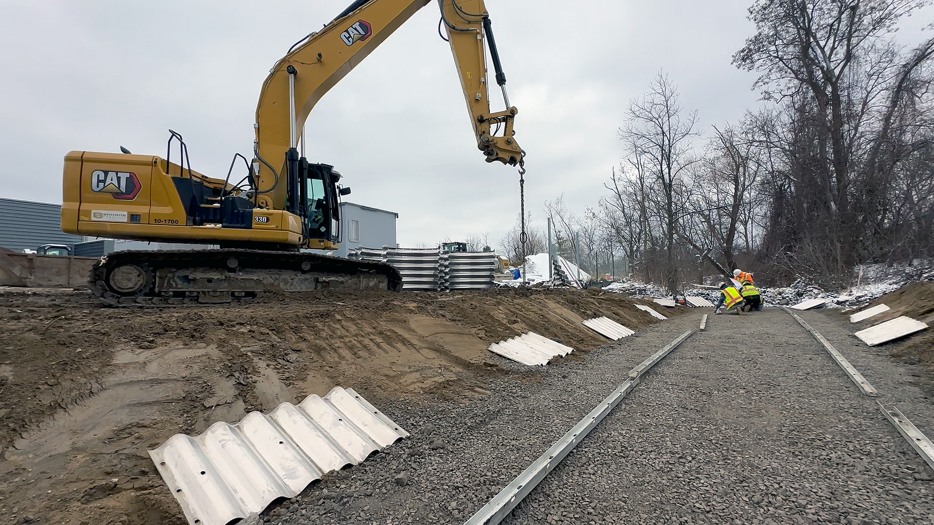 Underground utility construction workers installing railway tracks to connect large piping for underground utilities with a yellow excavator on a compacted gravel bed surrounded by trees.