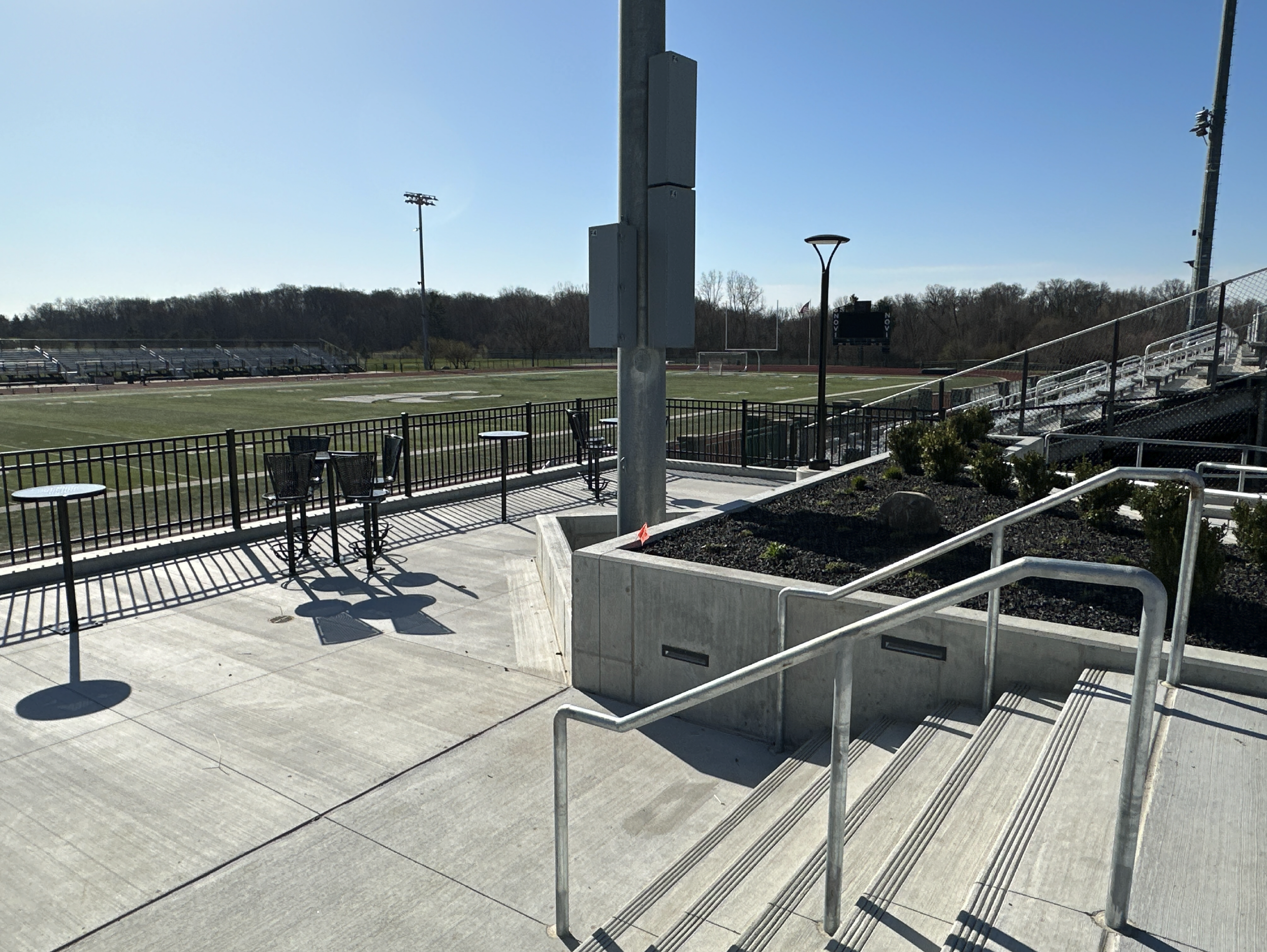 Empty sports stadium with finished concrete steps, pathways and vertical retaining walls with a football field, bleachers, and surrounding track, seen on a clear, sunny day at Novi High School.