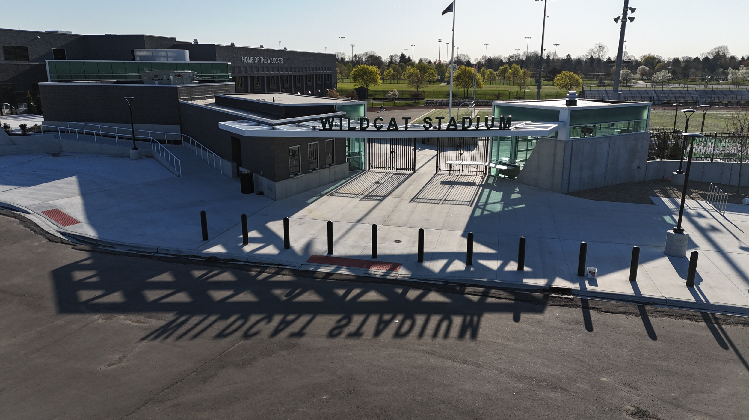 Entrance to Wildcat Stadium with a modern building and trees in the background, featuring a sign that says 'HOME OF THE WILDCATS' and a large shadow cast on the ground that reads 'WILDCAT STADIUM'.