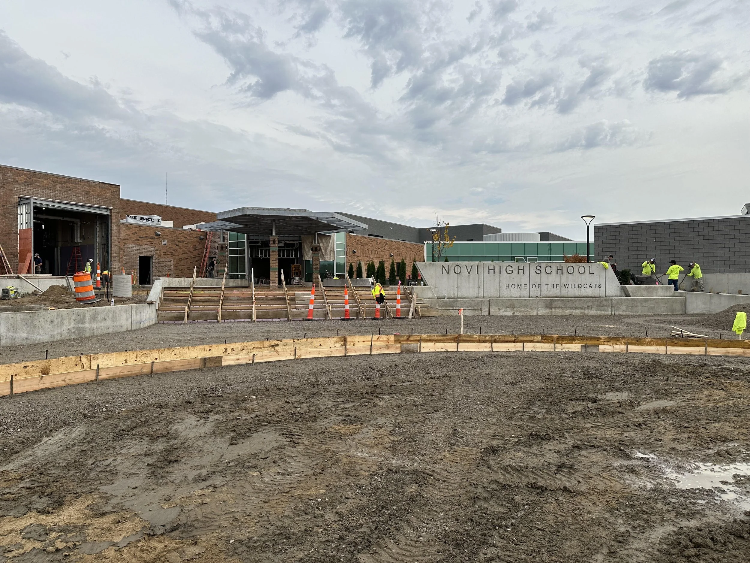 Concrete construction workers placing forms at the new Novi High School fitness center expansion with the sign 'Home of the Wildcats' visible in the front. The area is under construction, with dirt, wooden forms, and construction equipment present.