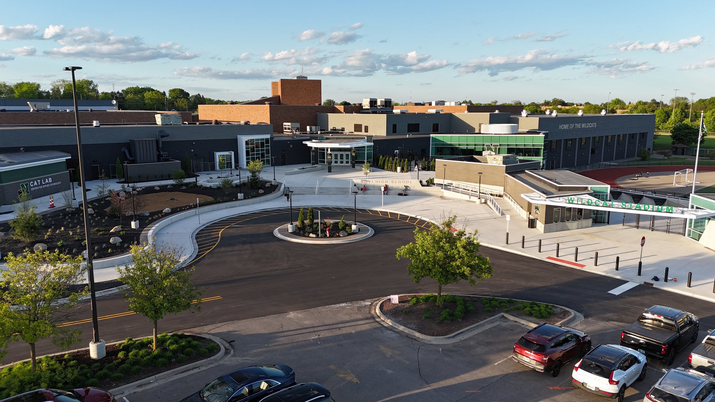 A high school campus with modern buildings labeled 'Novi High School', green signage for 'Wildcat Stadium', a parking lot with cars, a landscaped area with trees, and an outdoor basketball court under a partly cloudy sky.