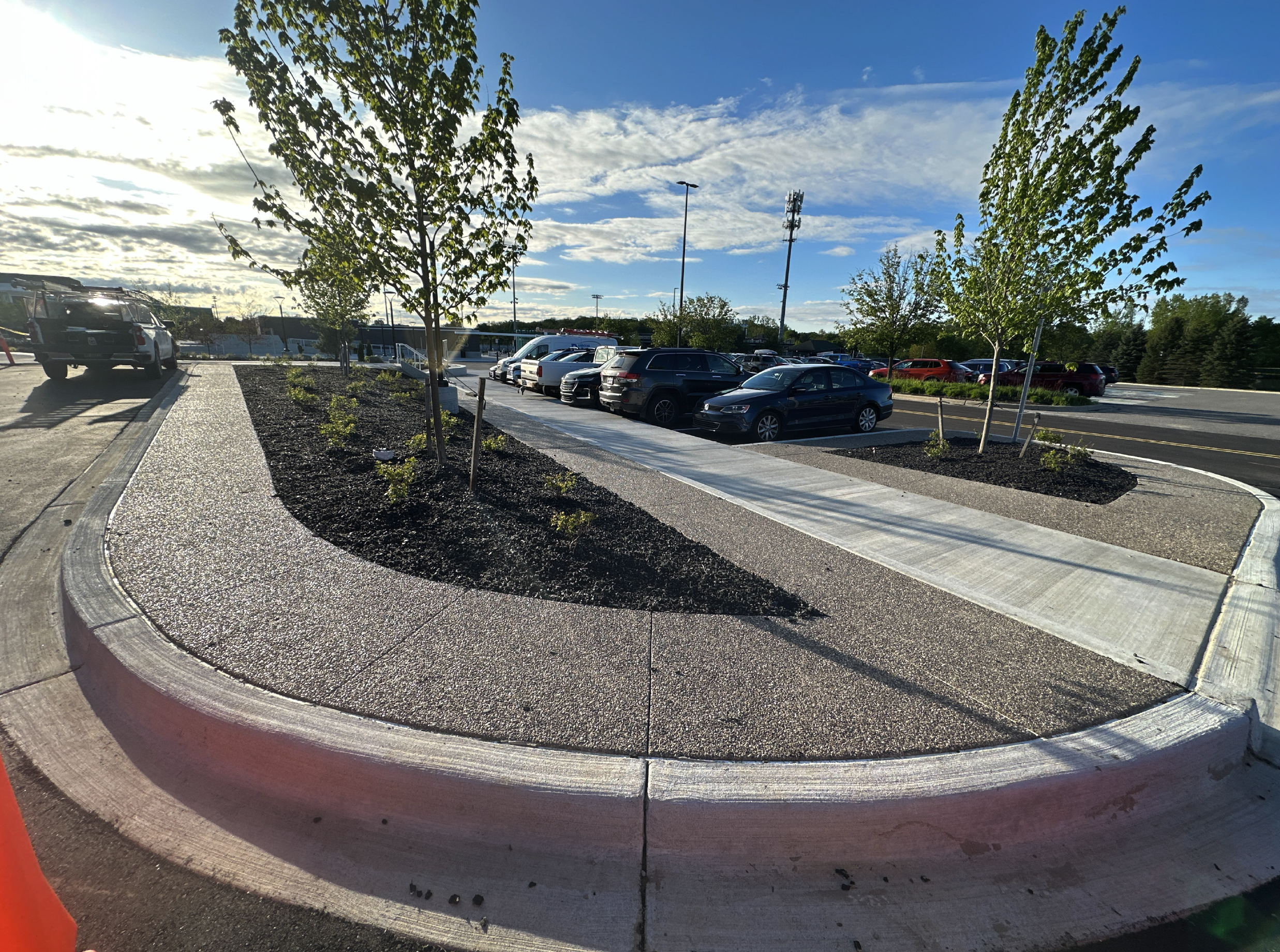 A parking lot with a sidewalk and landscaped area with trees and small plants, under a sky with clouds and sunlight.