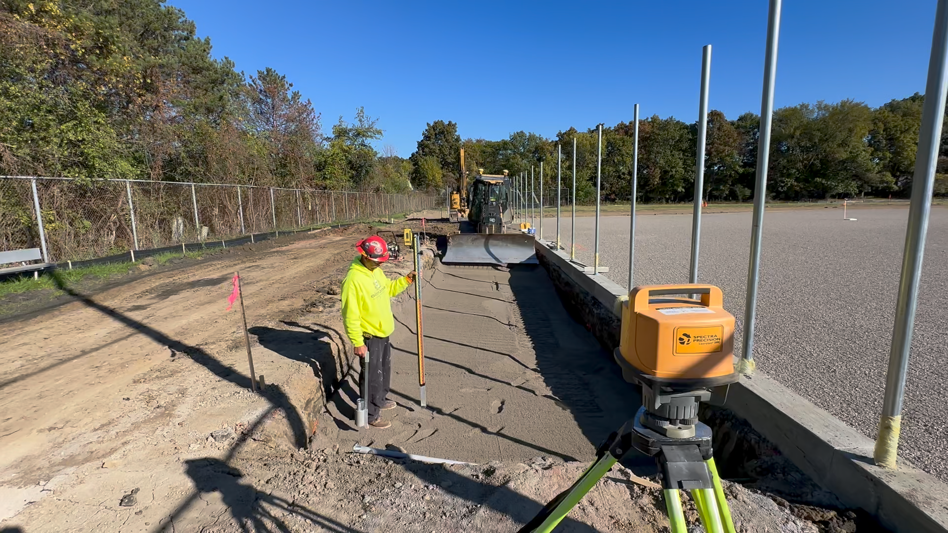 Earthwork construction worker in a bright yellow vest and red helmet operating lazer guided leveling equipment on a sports field construction site for Grosse Ile High School with a backhoe and trees in the background.