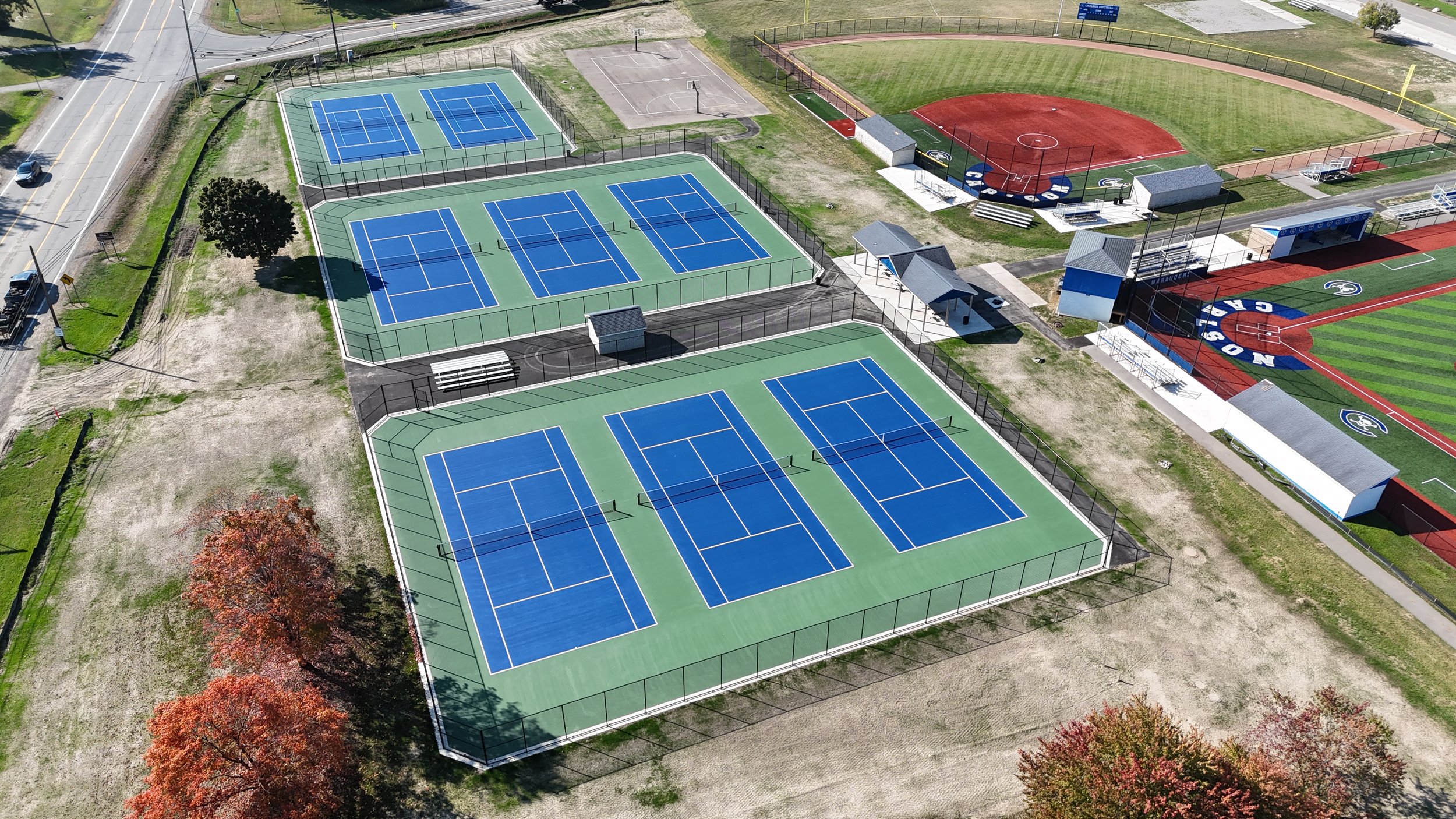 An aerial view of a sports complex showing multiple tennis courts, with some enclosed in fencing, and a baseball field with a track around it. The area includes small structures and sports equipment, and the surrounding area has trees with autumn foliage.