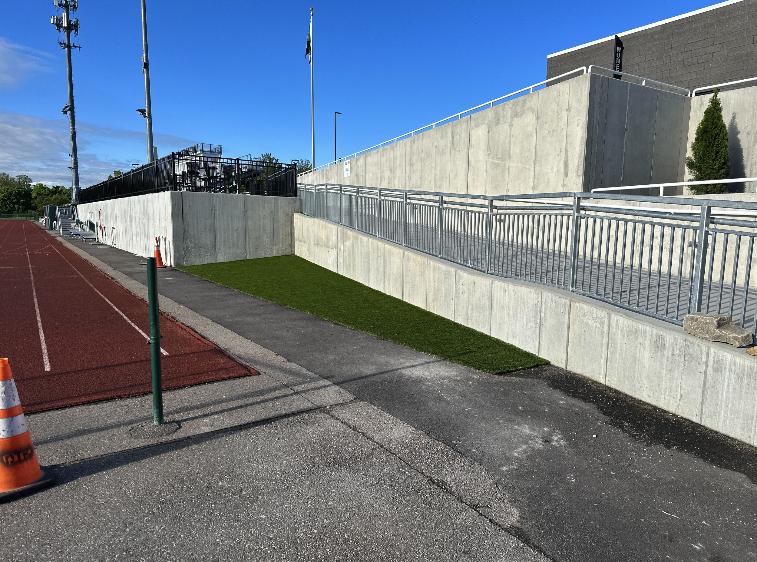 A running track adjacent to a concrete building with metal railings and a small patch of artificial grass, under a clear blue sky.