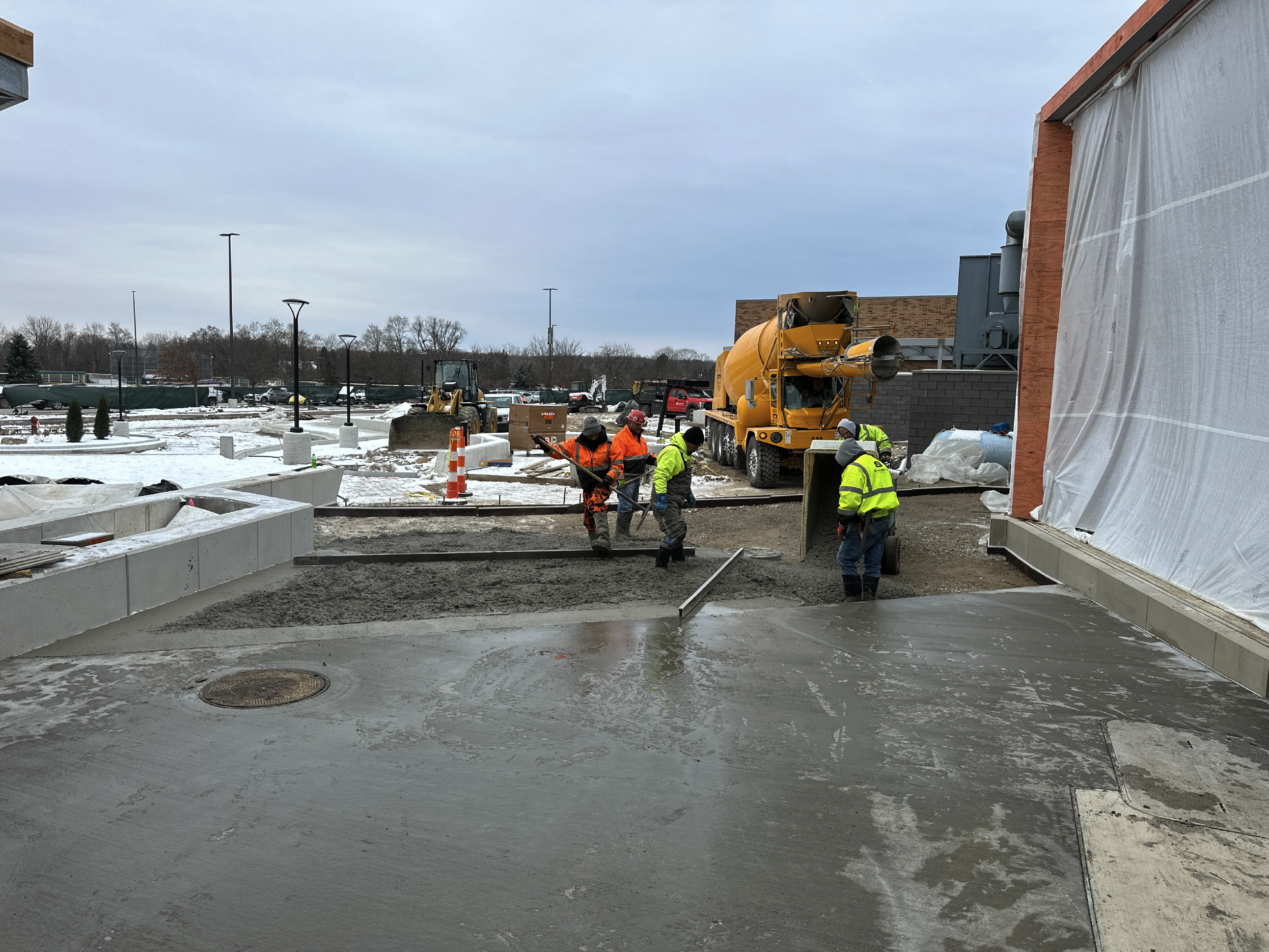 Construction workers pouring concrete outside a building on a snowy day, with construction equipment and materials nearby.