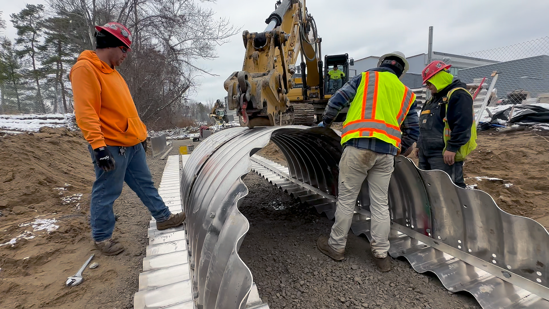 Underground utility construction workers using heavy machinery to install large, corrugated metal drainage pipes Troy Smith Middle School project construction site.