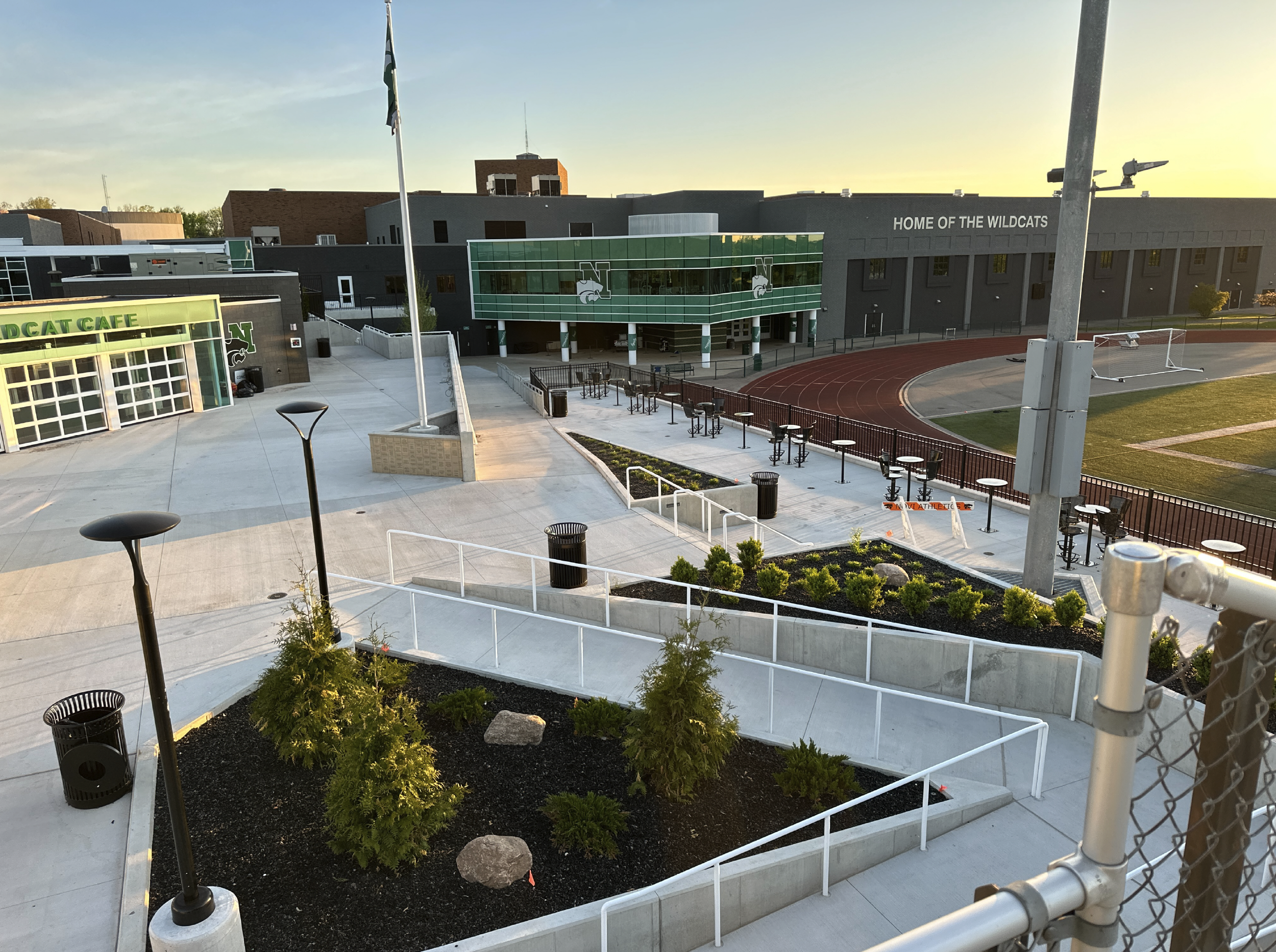 A school campus with a track and field area, a sports field, and modern school buildings. The school is named 'Wildcats' and has a green and gray color scheme. The area is clean and well-maintained with landscaped plants and outdoor seating.