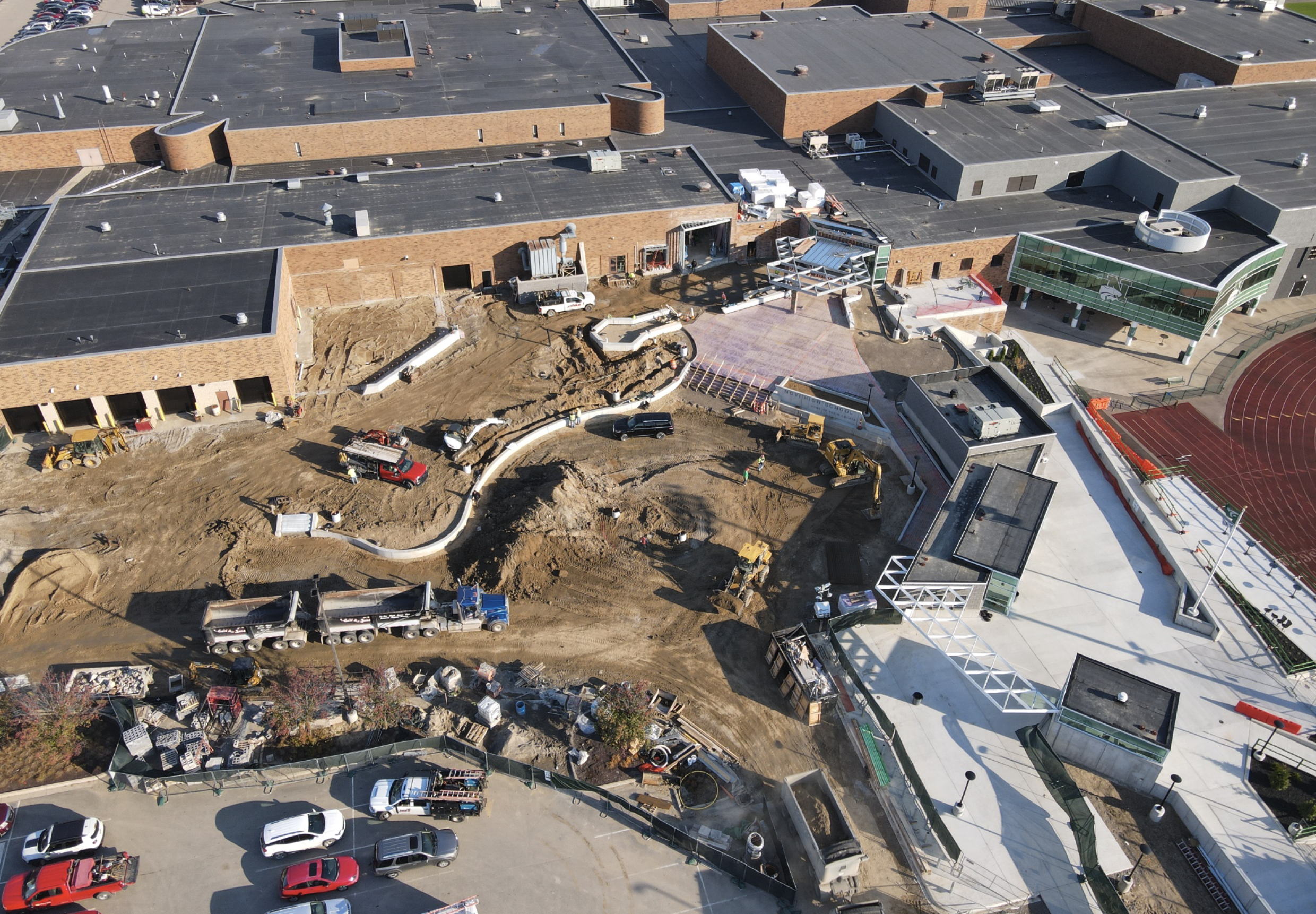 Aerial view of a school campus under construction with building structures, construction vehicles, and workers, including a track field on the right side.