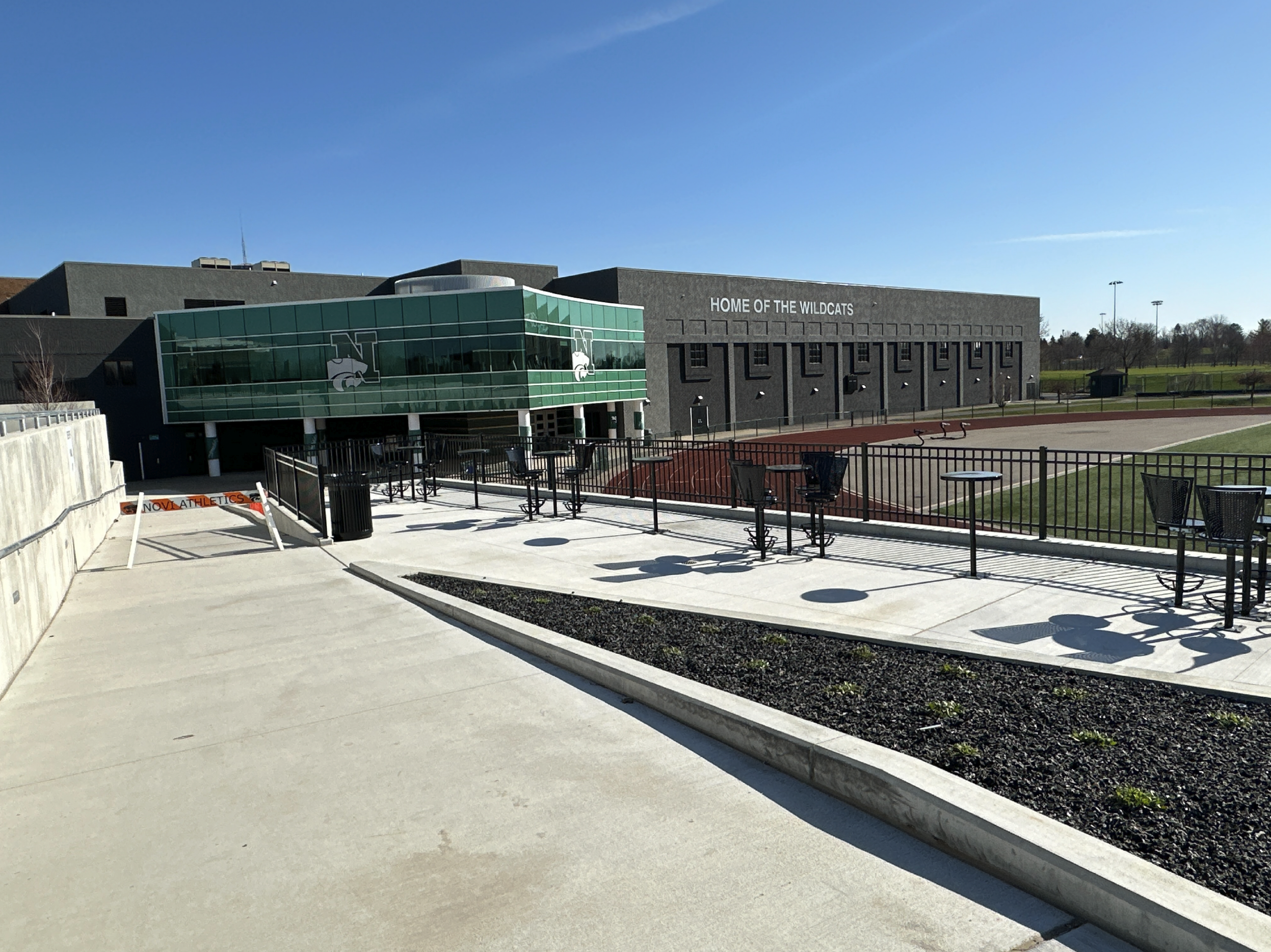 A school athletic facility with a modern building featuring large green glass windows and a logo of a wildcat. There is an outdoor track surrounding a field with green and red artificial turf, and a small exercise area with bikes and chairs. The building has a sign that reads "Home of the Wildcats." The sky is clear and sunny.