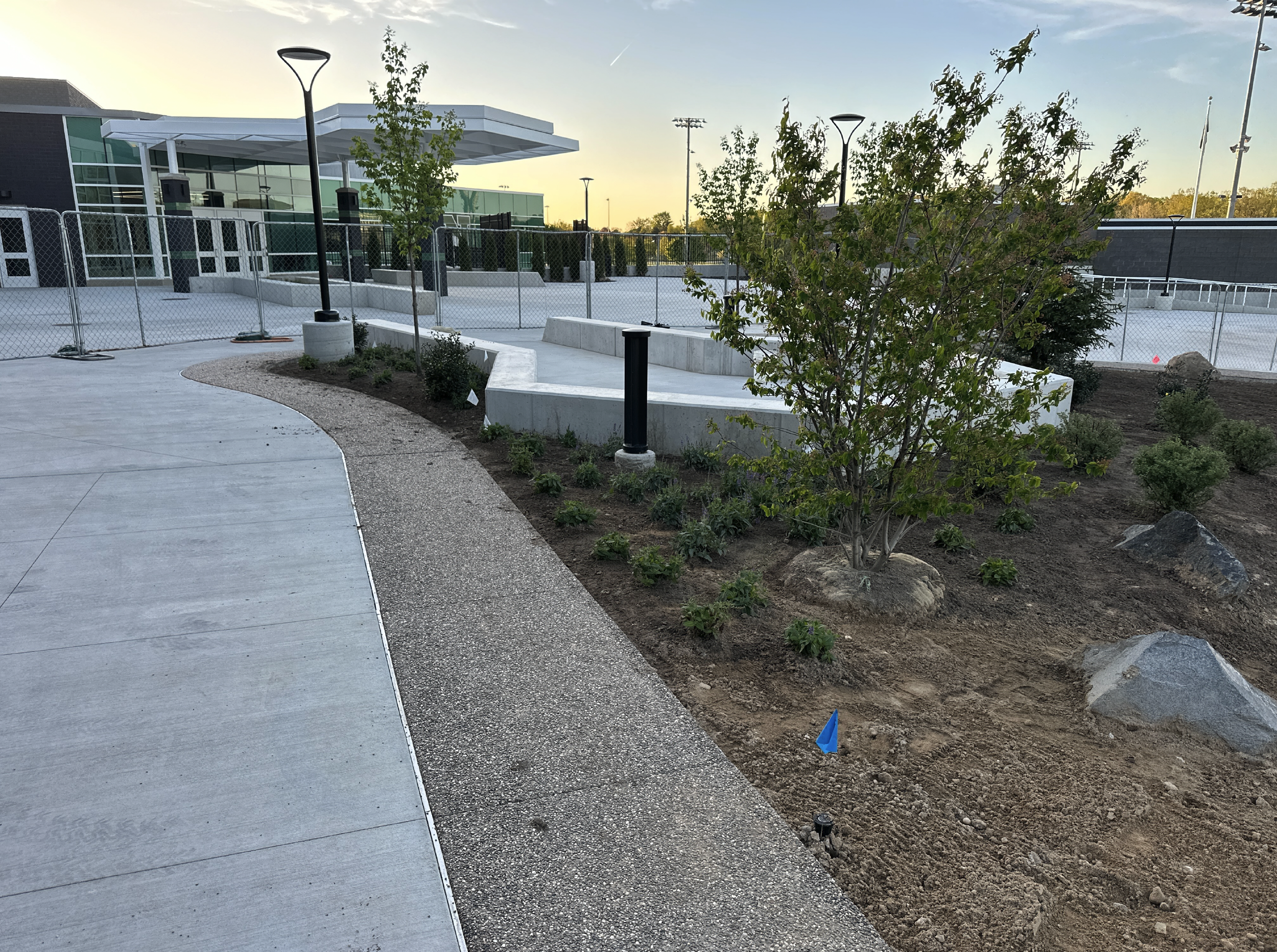Outdoor scene of a landscaped area with a concrete sidewalk, small trees, shrubs, rocks, and garden beds. There are modern street lamps, a building with glass windows in the background, and a chain-link fence surrounding the area. The sky is clear with the sun setting.