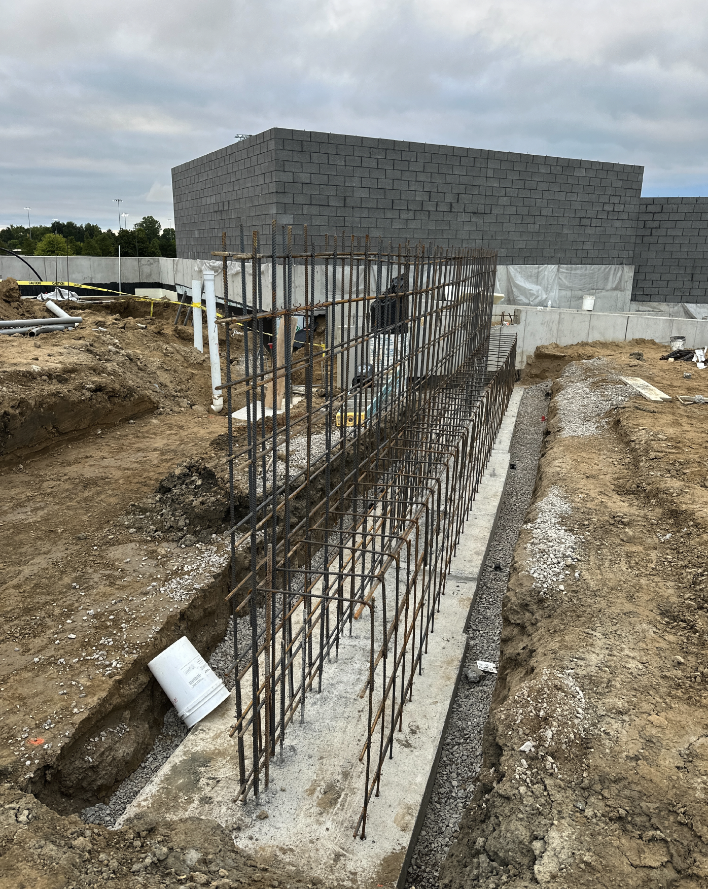 Construction site with rebar framework, dirt trenches, and a partially built concrete wall and foundation, with cloudy sky overhead.
