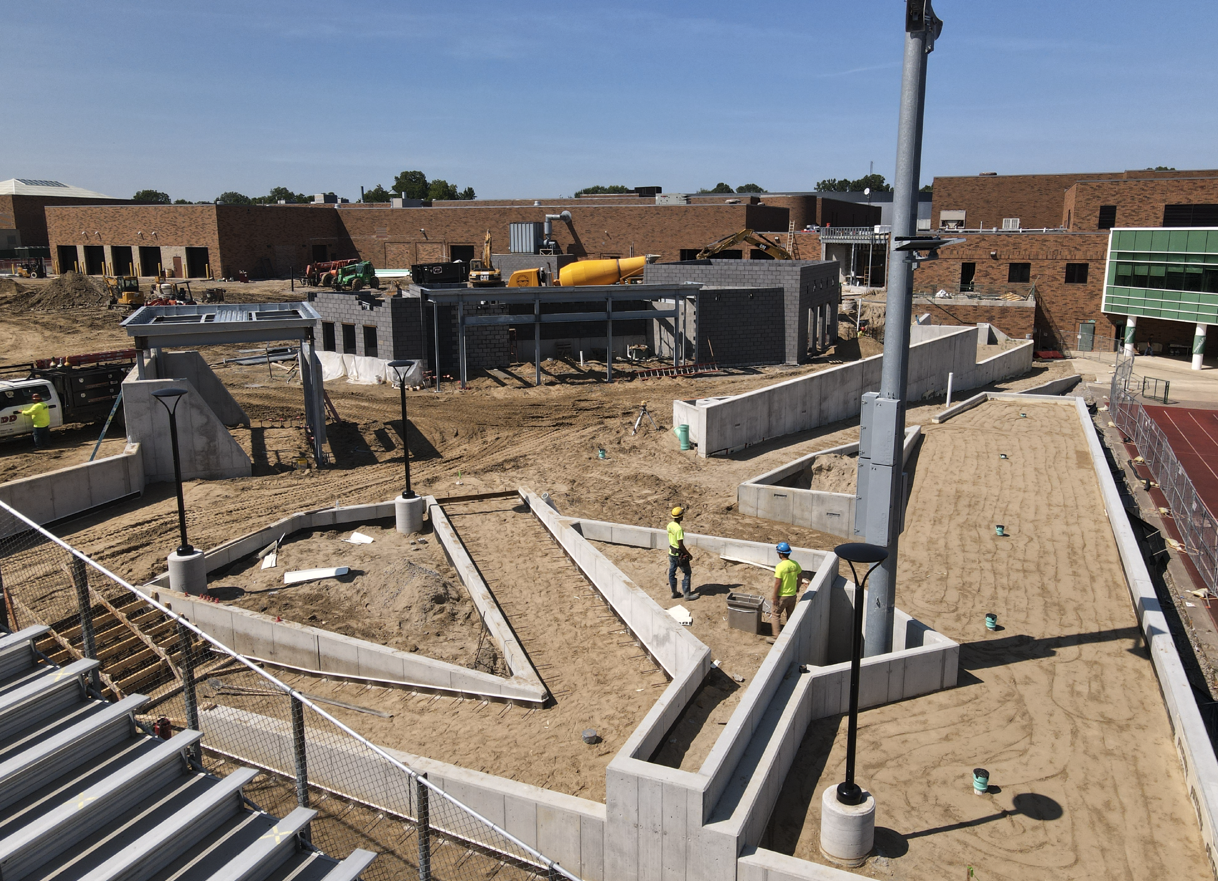 Construction site with workers, construction materials, and partially built structures, including concrete walls and a yellow cement mixer, on a sunny day.