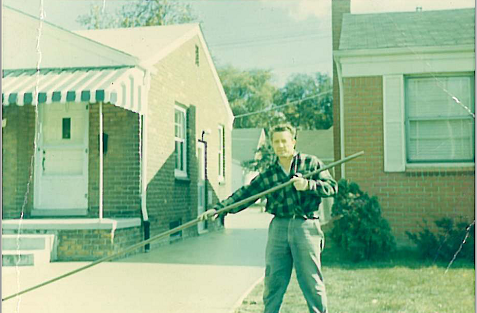 A man standing outside in front of brick buildings, holding a long stick or pole.