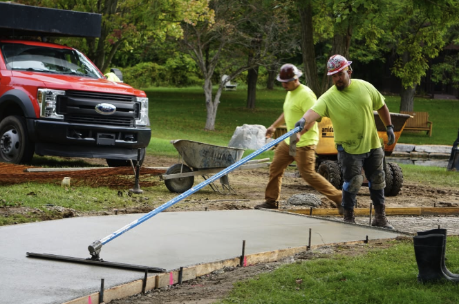 Two construction workers leveling freshly poured concrete on a sidewalk using a long screed tool, with a red pickup truck and trees in the background.