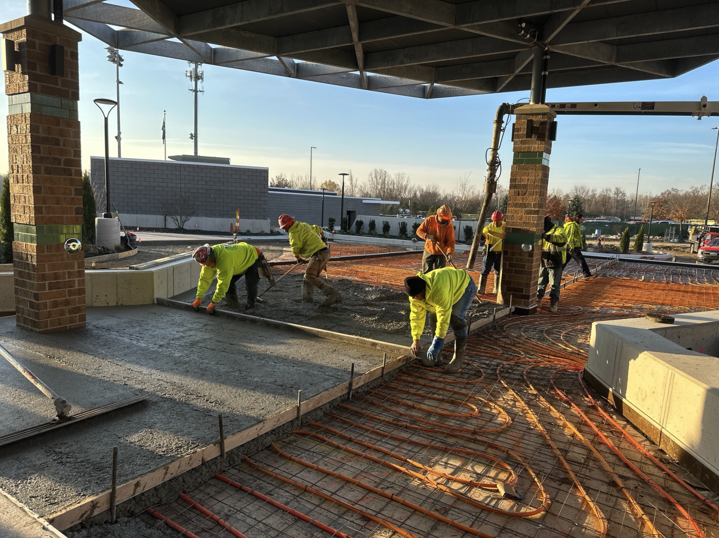 Construction workers in high-visibility clothing and hard hats pouring and leveling concrete on a building site with exposed orange heating cables and framing for a sidewalk or floor.