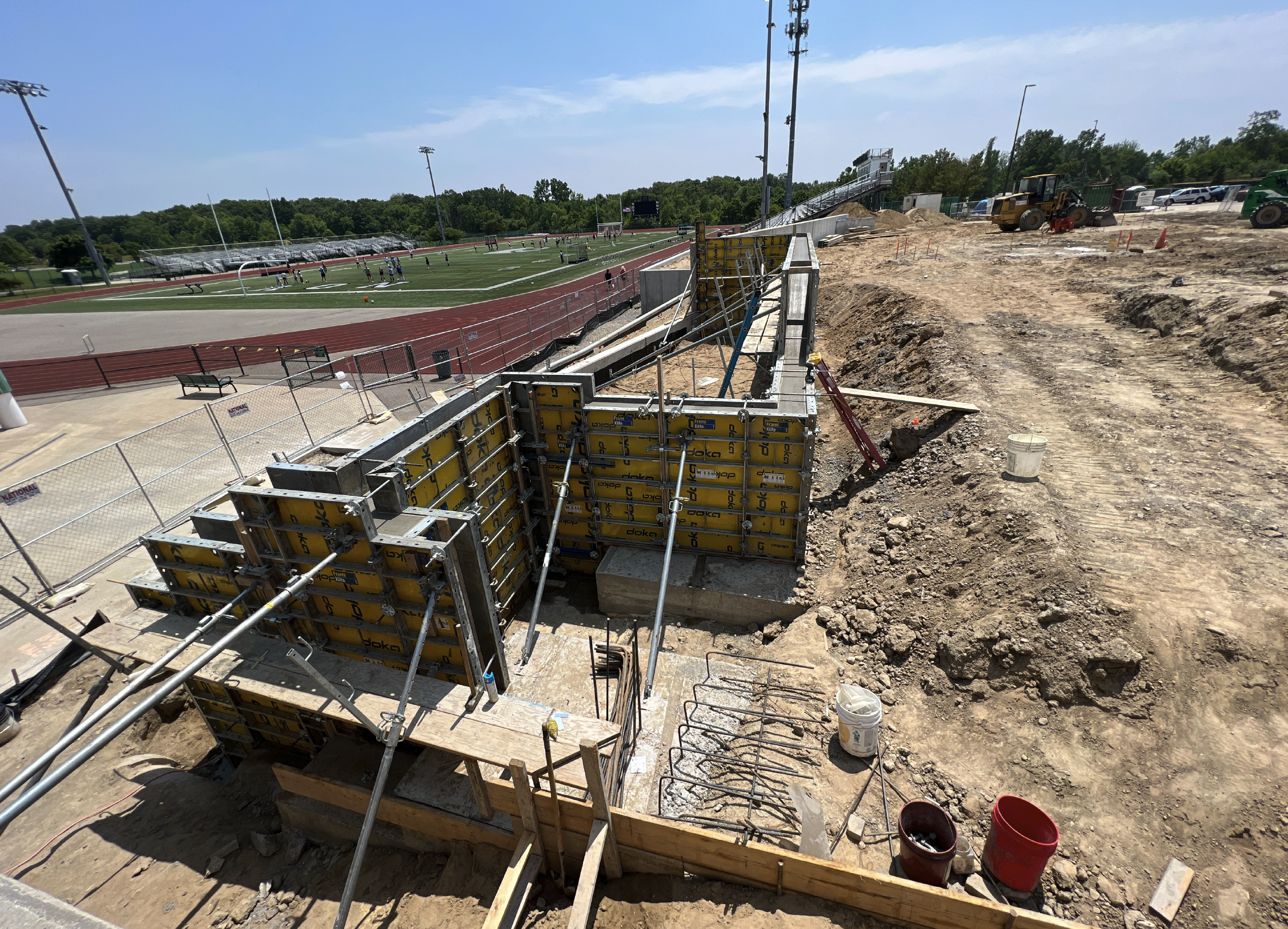 Construction site with concrete formwork and rebar near a sports stadium with a running track and football fields in the background.