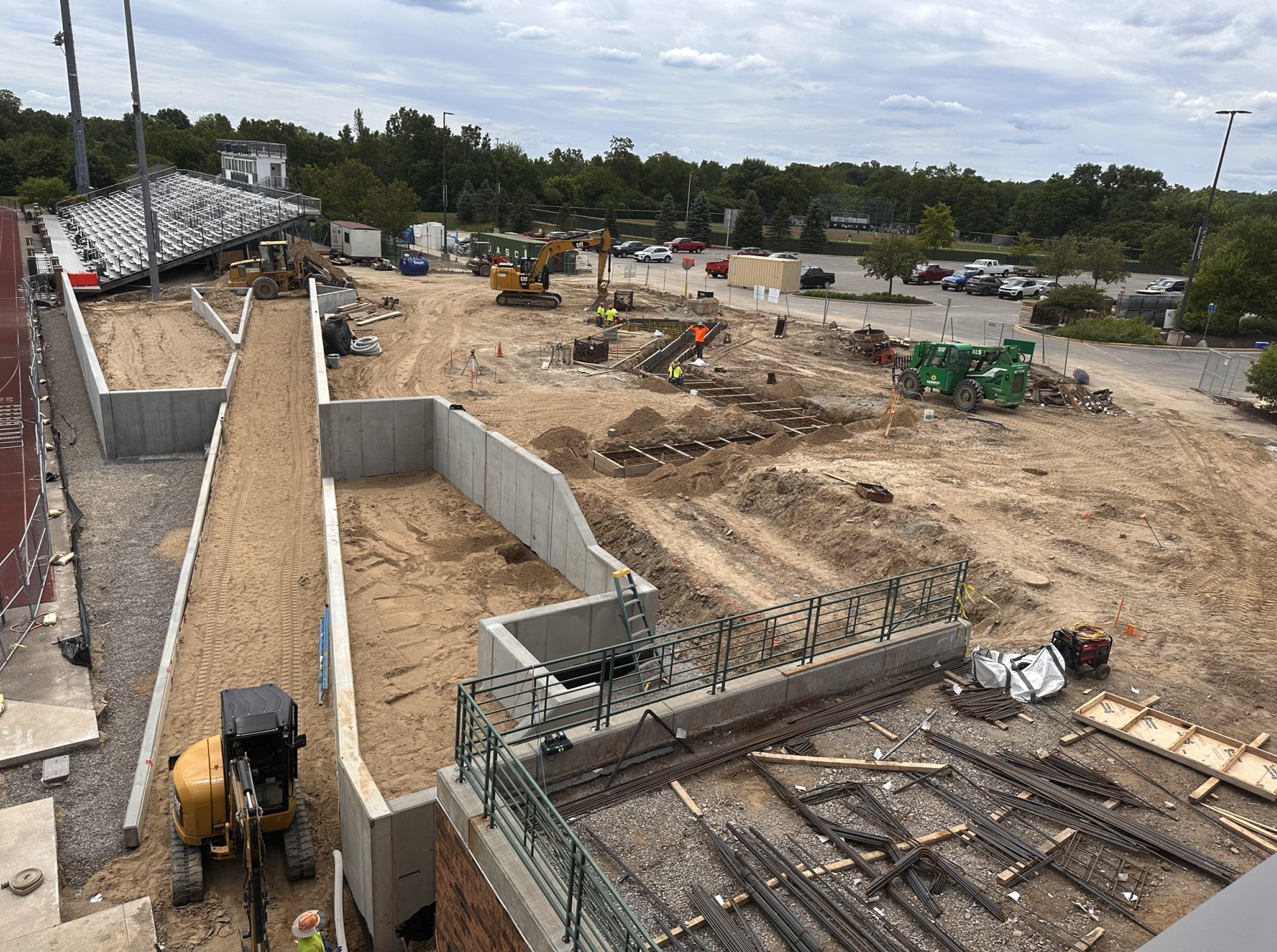 Construction site with workers, machinery, and tracks for future building or infrastructure, with a parking lot and trees in the background.