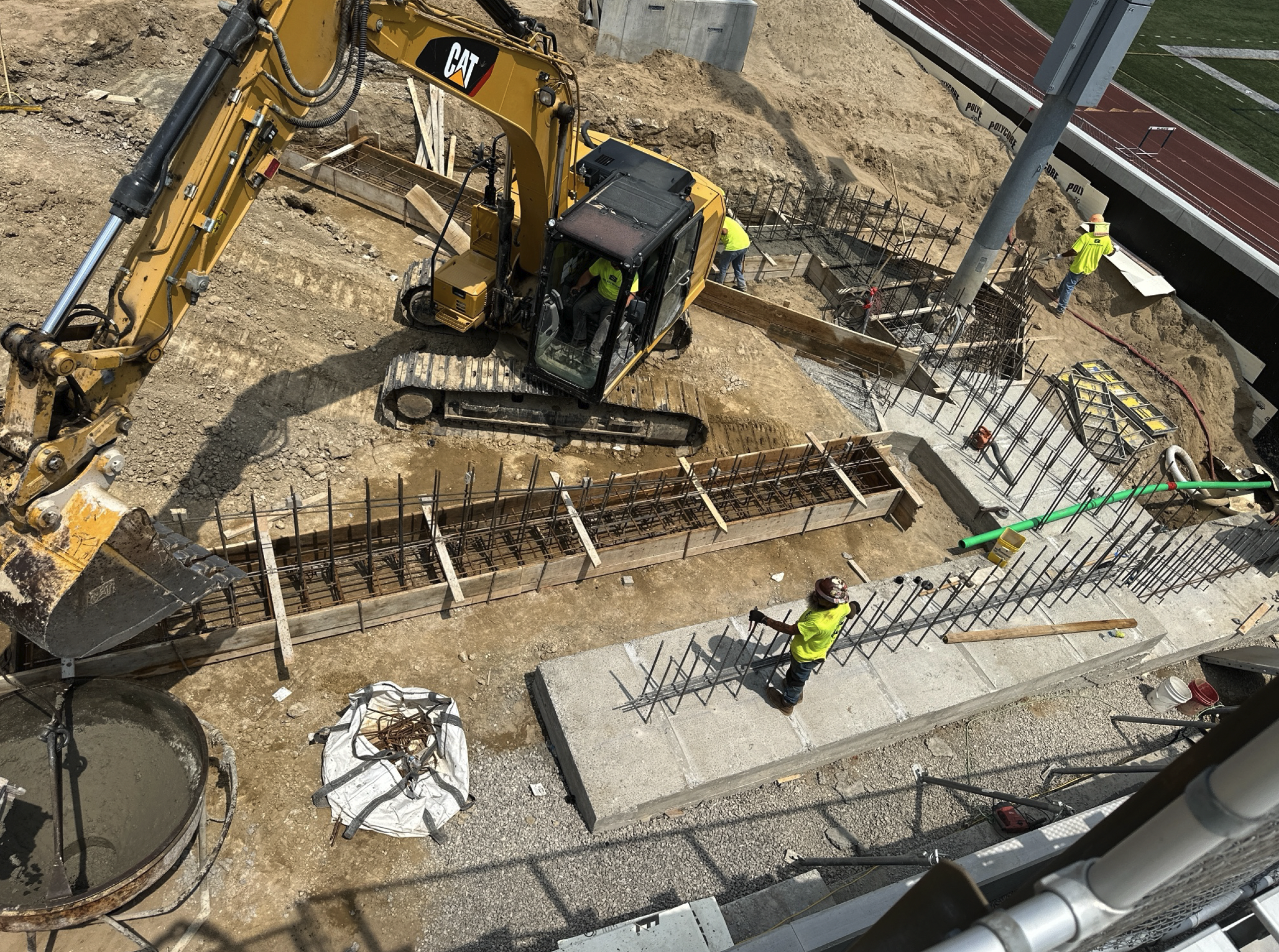 Construction site with workers and heavy machinery, building a foundation or structure with rebar and concrete. Green pipe protrudes from the ground, with a track and field in the background.