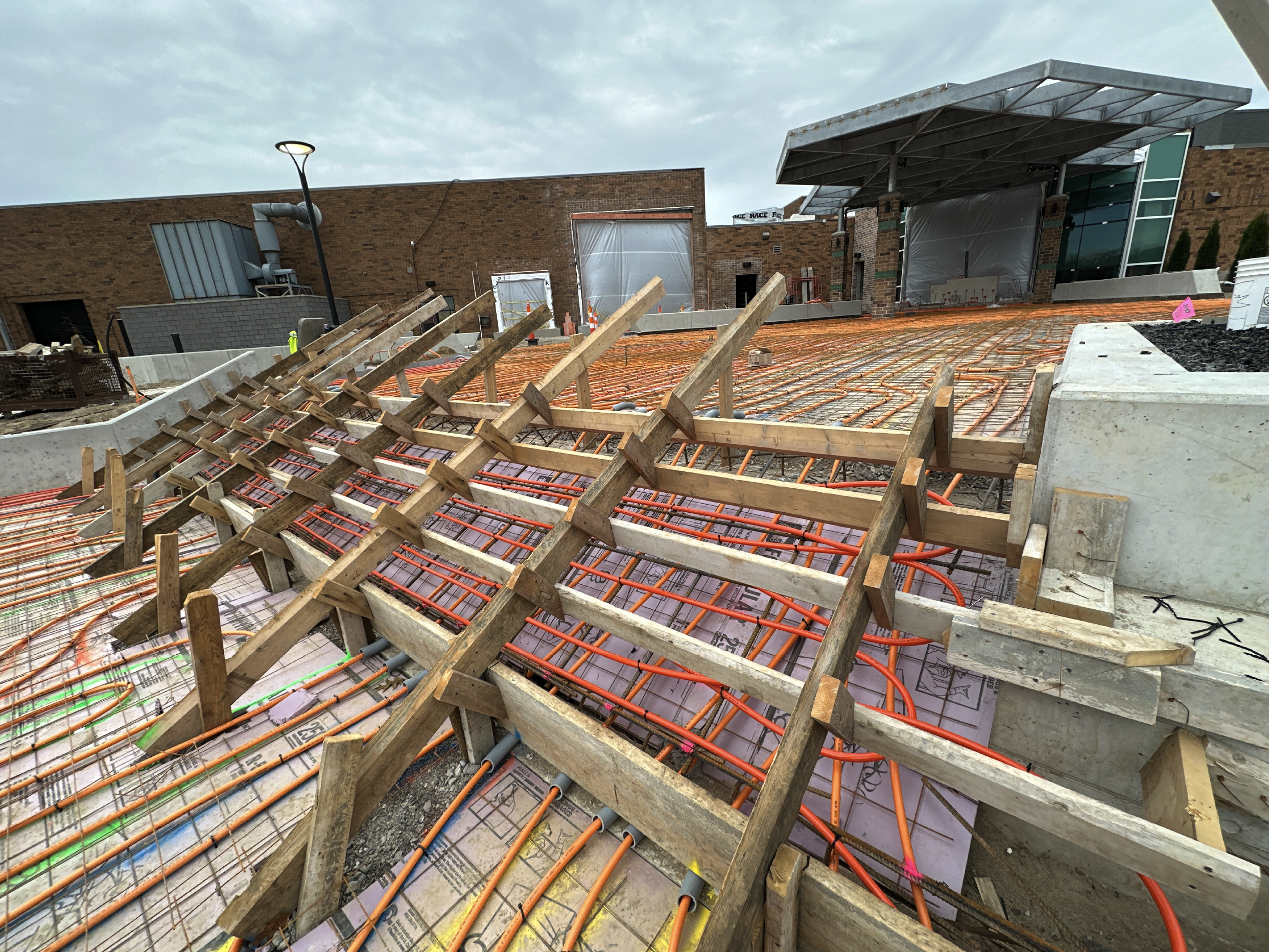 Construction site with orange heated floor pipes and wooden formwork, with a building structure in the background.