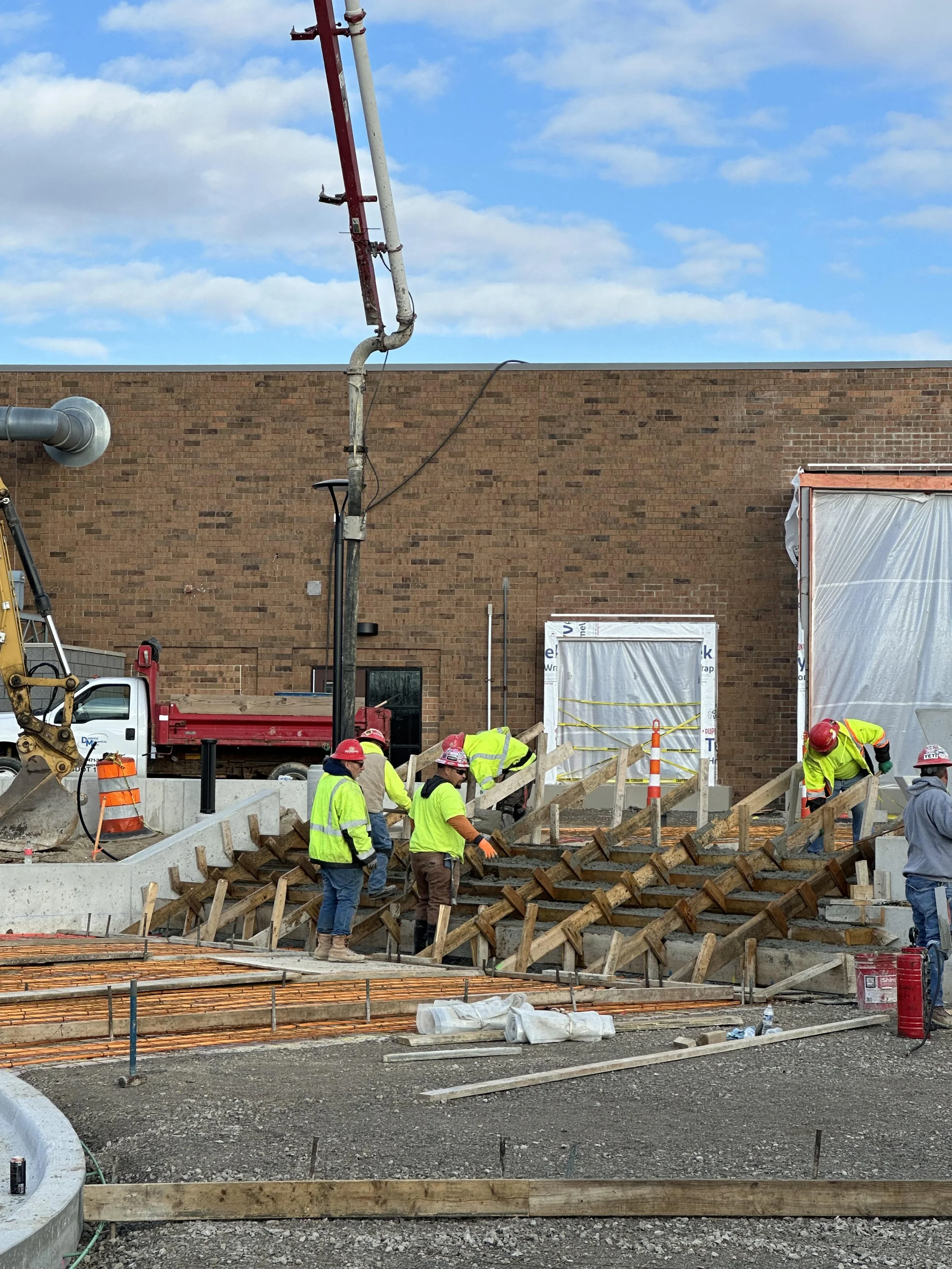 Concrete construction workers wearing red Simone Companies hard hats building a staircase at the new Novi High School fitness center expansion. Construction materials, concrete pump and tools are visible around them.