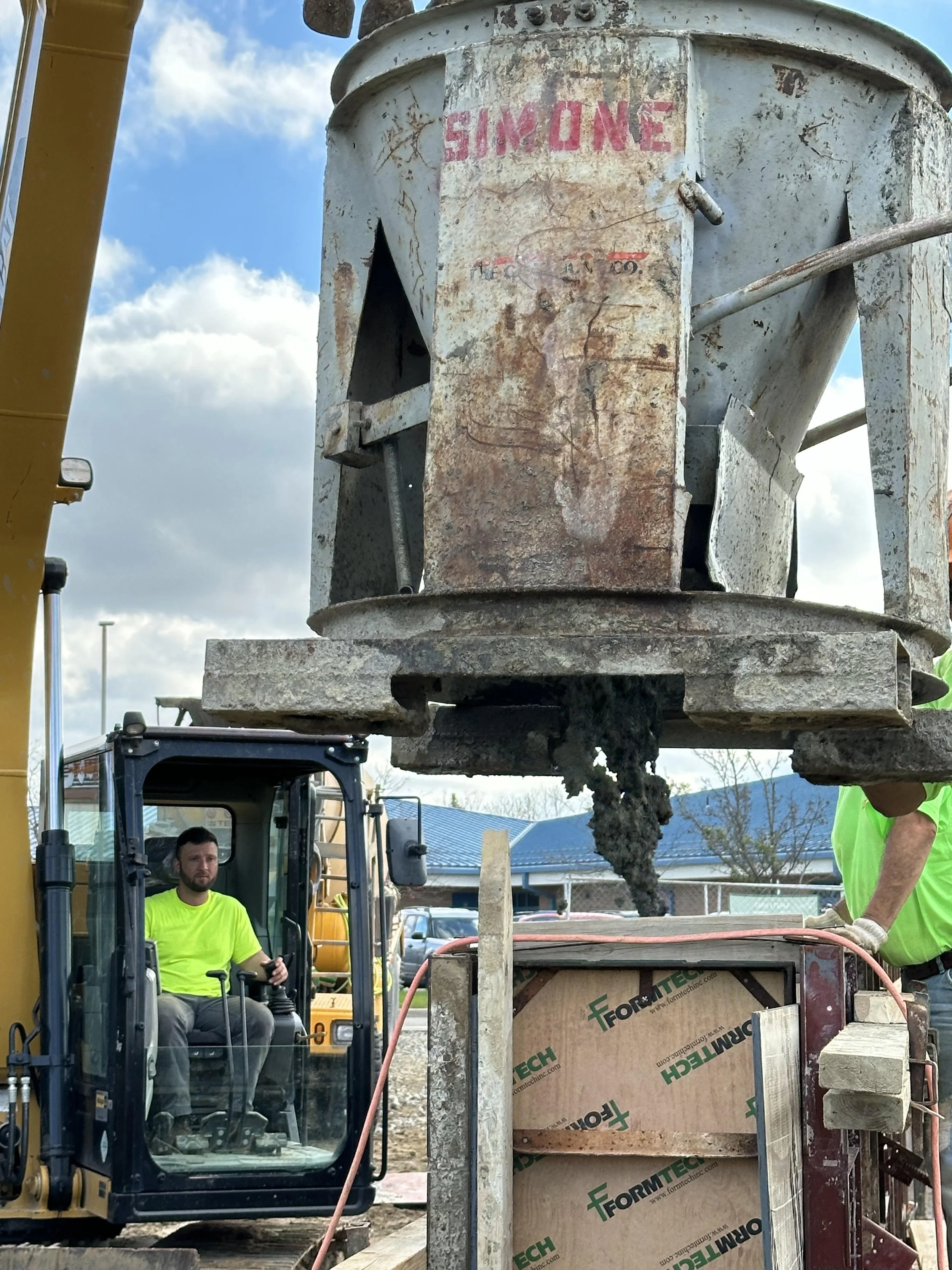 Construction workers operating a concrete pump to pour concrete into a foundation or section of a building. One worker is seated in a yellow excavator, and another is standing nearby.