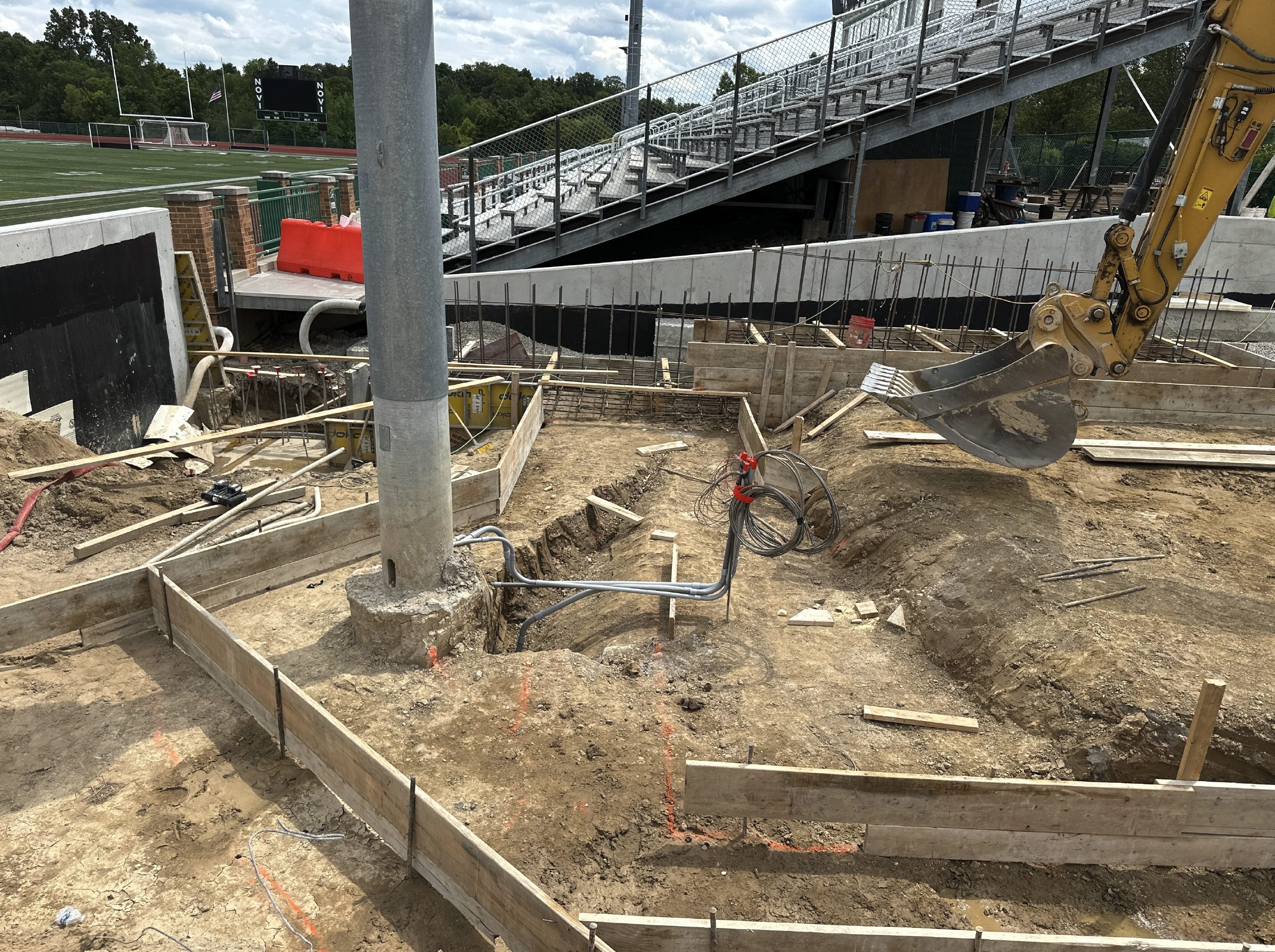 Construction site with dirt, wooden frames, electrical and plumbing wiring, and a small excavator with a bucket attachment, near a sports field with bleachers.
