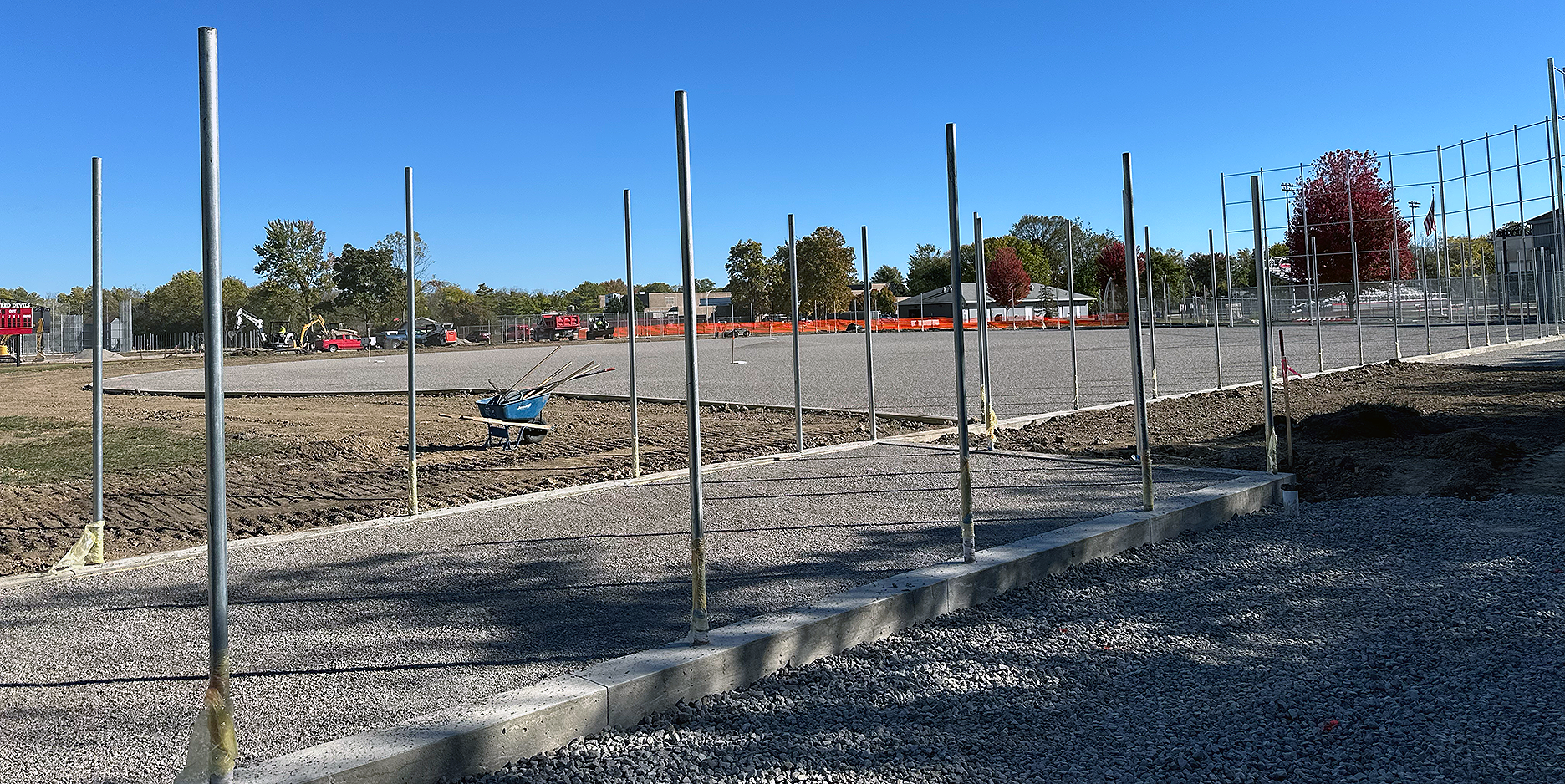 Construction site with a partially built sidewalk, metal posts, and a fenced sports field in the background on a sunny day.
