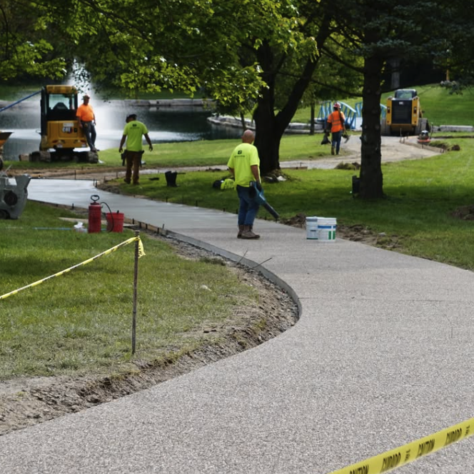 Workers in safety vests installing or repairing a walking path in a park near a pond, with construction equipment and caution tape present.