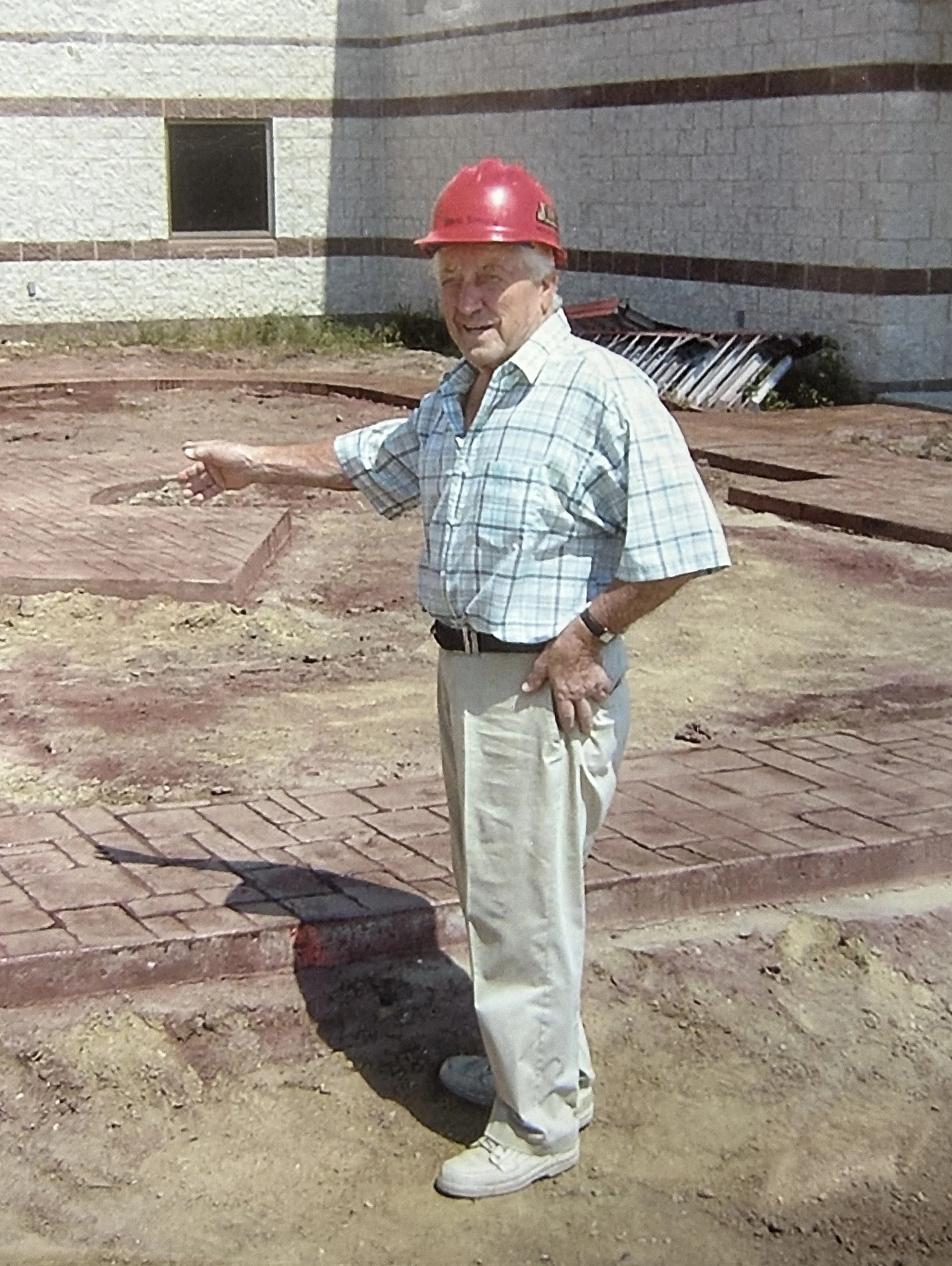 Older man wearing a red construction helmet and casual clothing pointing at a construction site with a brick sidewalk and a building in the background.