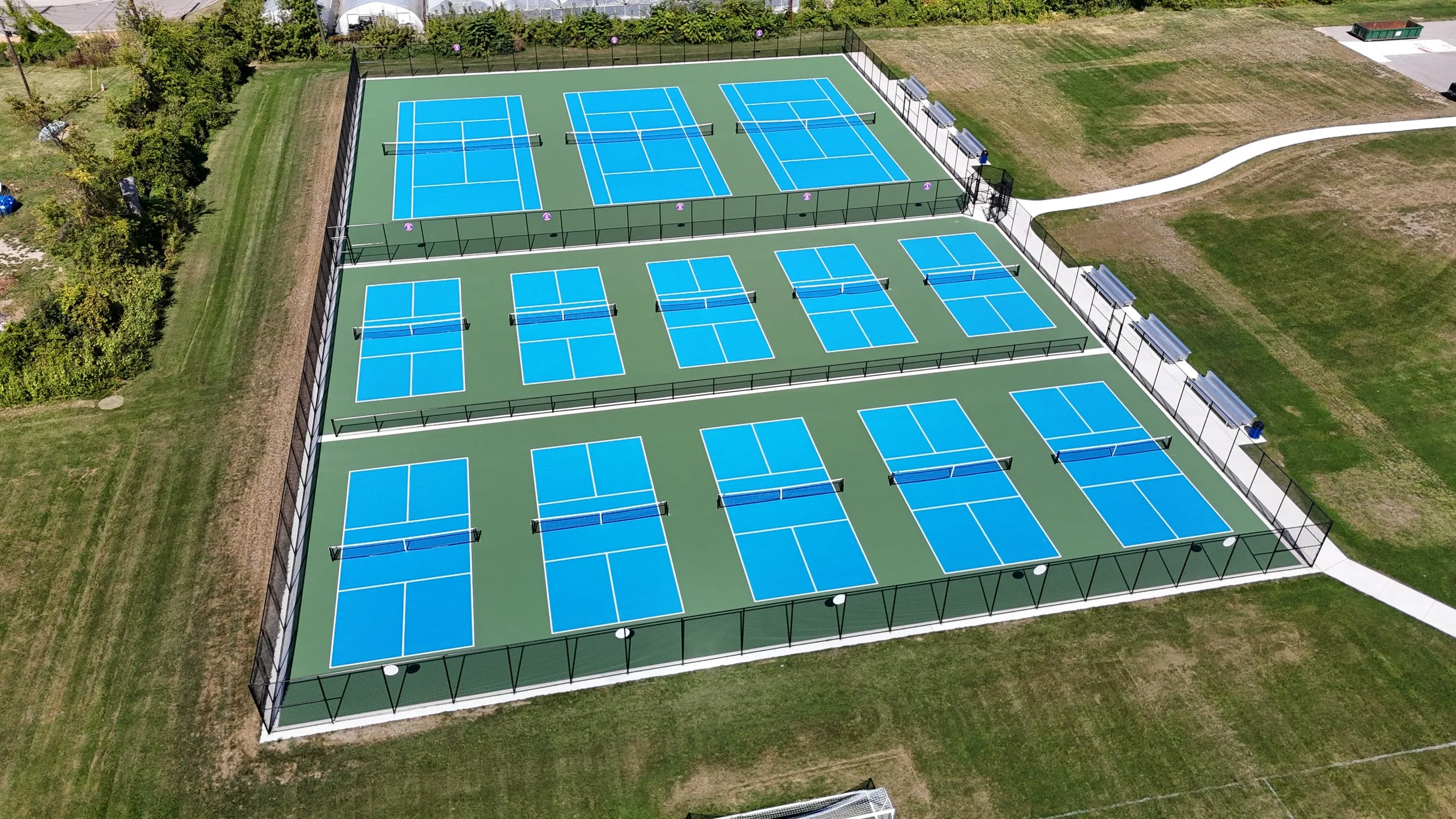 Aerial view of ten blue tennis courts with white lines, surrounded by a black fence, benches, and green grass.