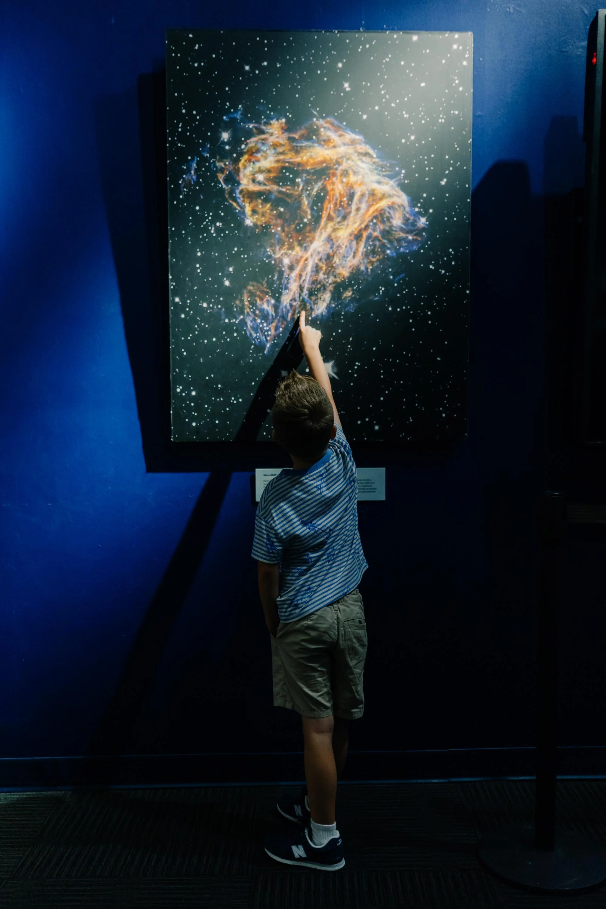 A young boy pointing at a large canvas of a galaxy or nebula at a science museum or exhibit, with a dark blue wall in the background.