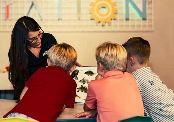 A woman engaging with three children at a table, showing them a book or educational material, with a colorful educational chart in the background.