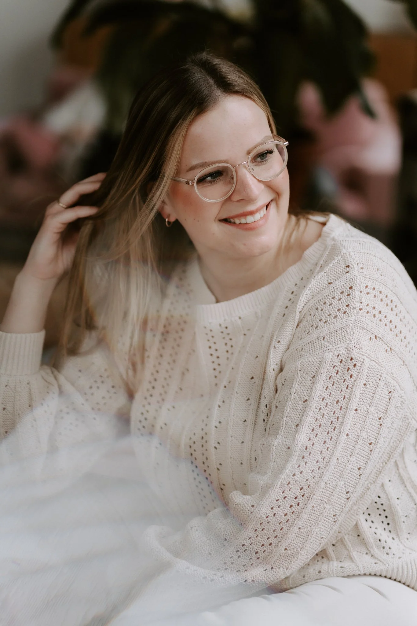 Junge Frau mit Brille und weißen Pullover lächelt, während sie in einem gemütlichen Raum sitzt.
