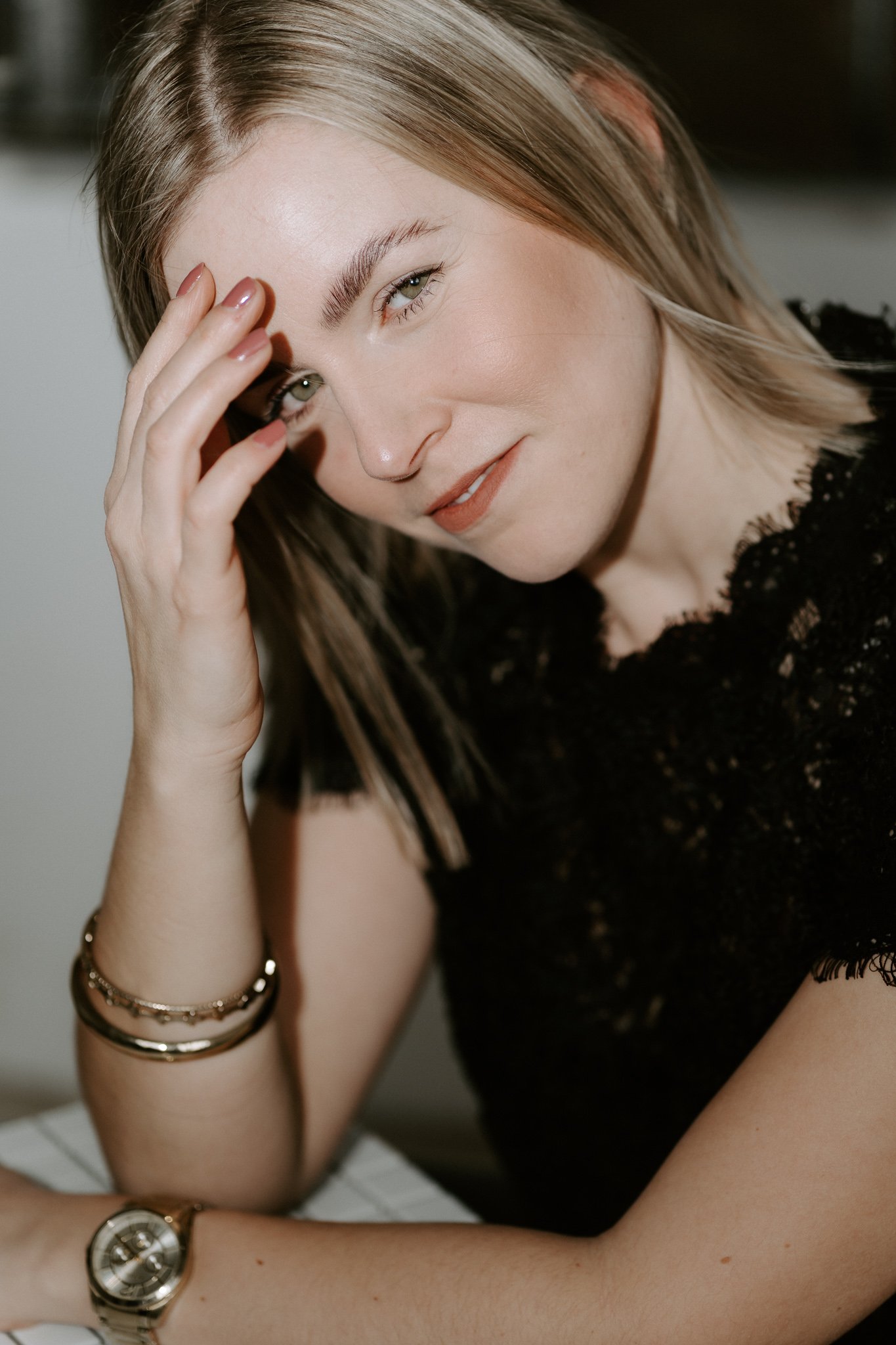 Nahaufnahme einer lächelnden Frau mit Blonden Haaren, die die Hand an den Kopf hält, trägt eine schwarze Spitze Bluse, Armband und Uhr, vor hellem Hintergrund.