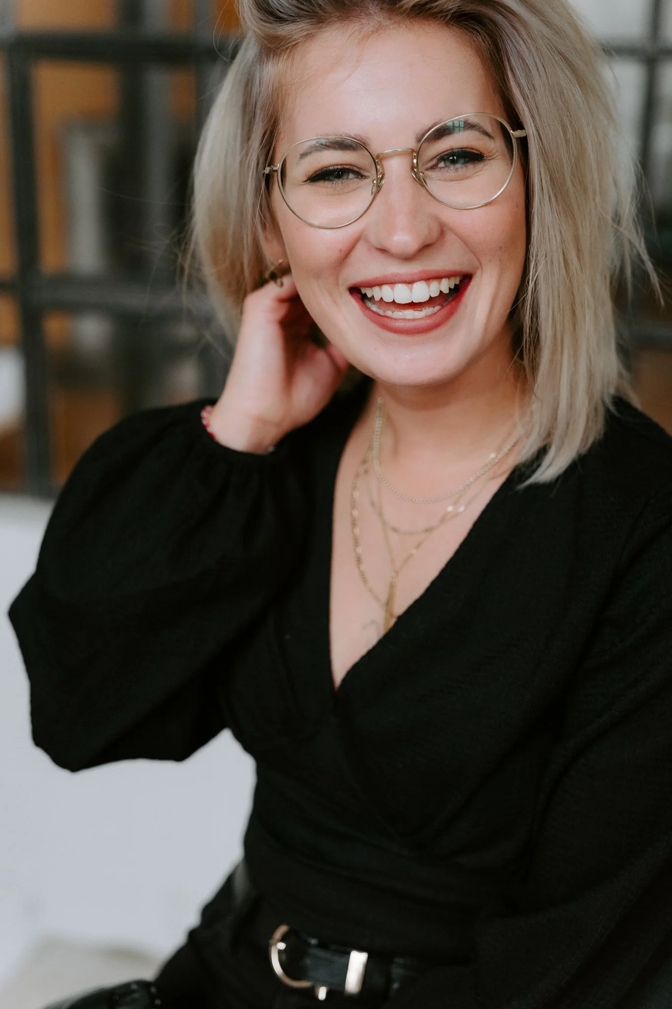 Lächelnde junge Frau mit Brille und blonden, gestylten Haaren, trägt schwarze Kleidung und mehrere Halsketten, im Hintergrund eine moderne, industrielle Inneneinrichtung.