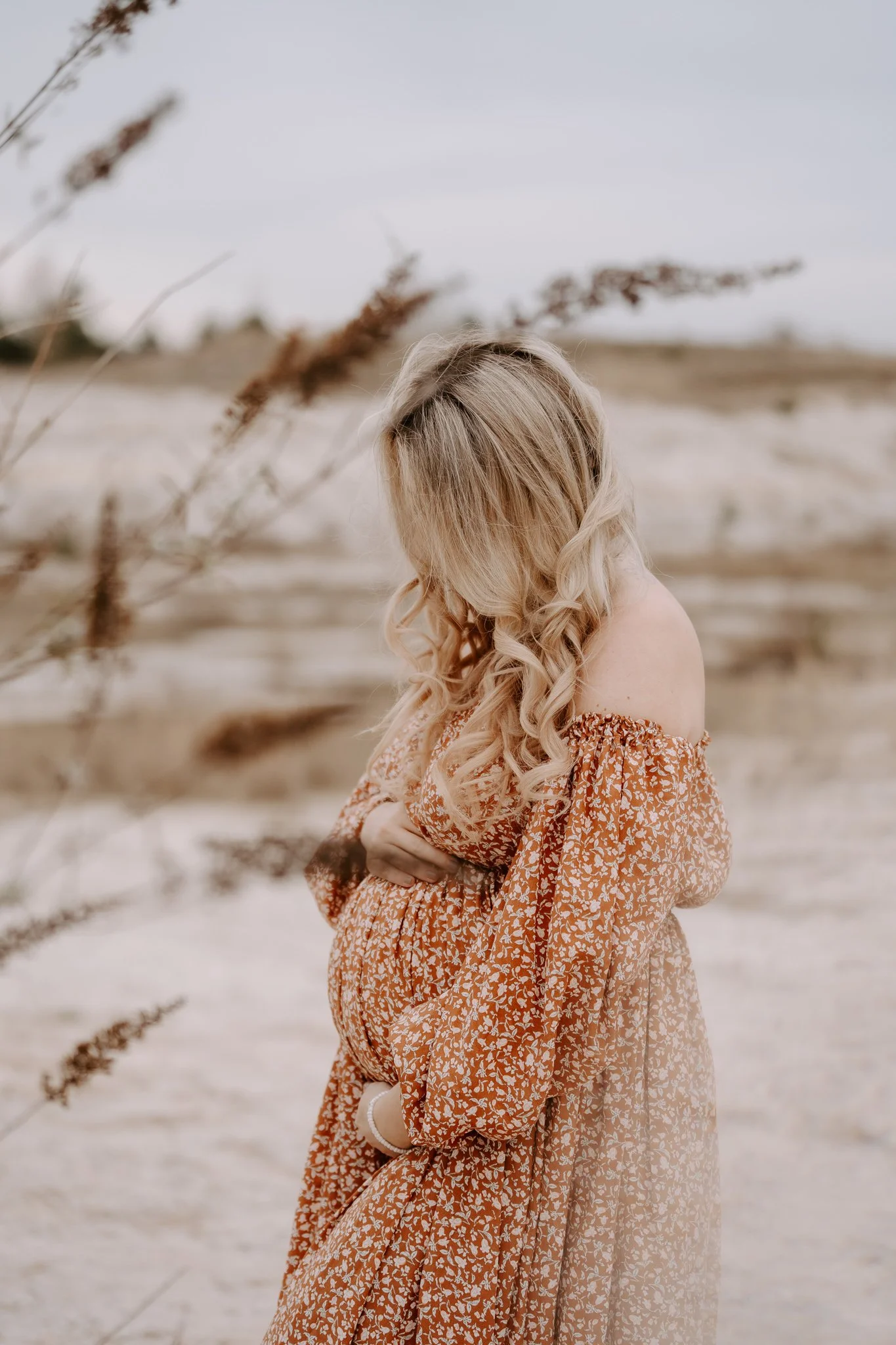 Frau mit langem, gewelltem Haar in einem orangefarbenen, gemusterten Kleid steht an einem Strand, den Blick nach unten auf ihren Babybauch gerichtet.