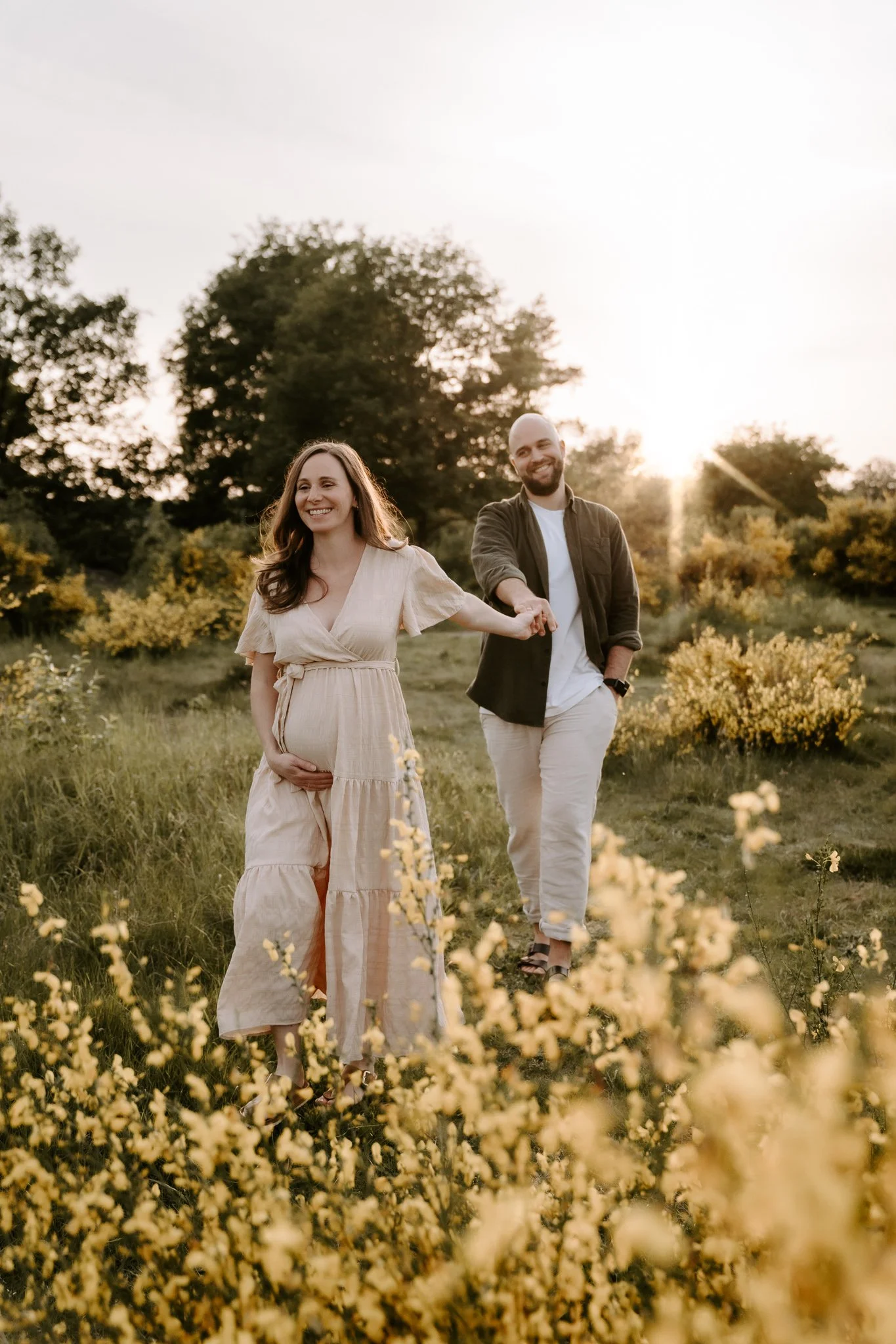 Eine schwangere Frau und ein Mann halten sich an den Händen, spazieren durch ein Feld mit gelben Blumen bei Sonnenuntergang.
