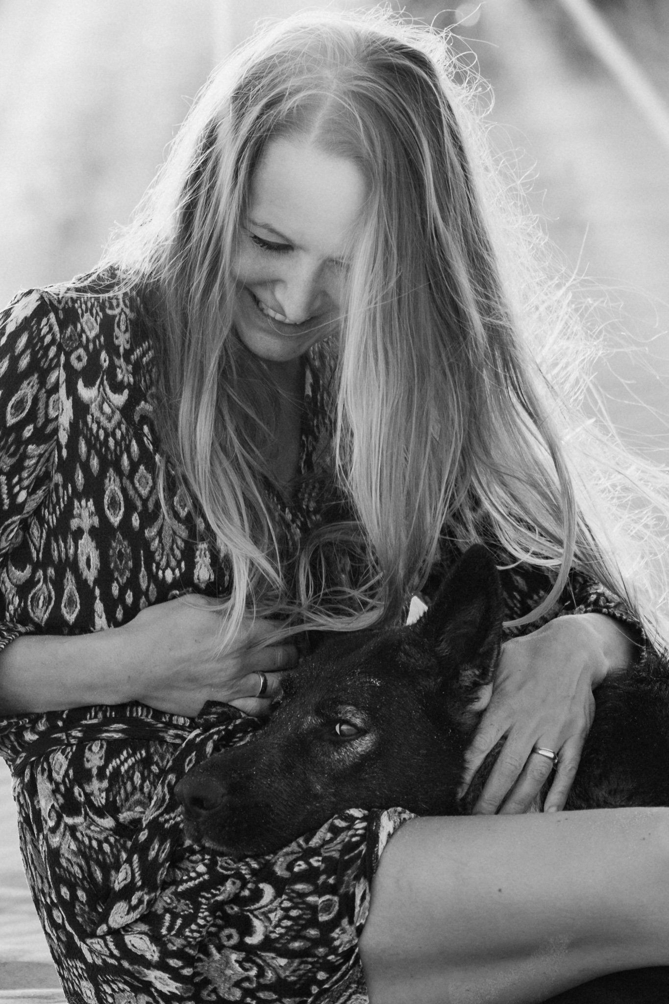 Eine lachende Frau mit langen, welligen Haaren, die einen Hund umarmt.