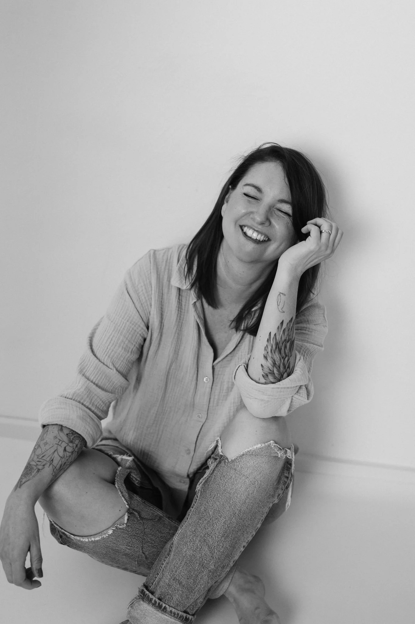 Schwarz-Weiß-Foto einer lachenden Frau mit Tattoos, sitzend gegen eine Wand, tragende Jeans und Hemd