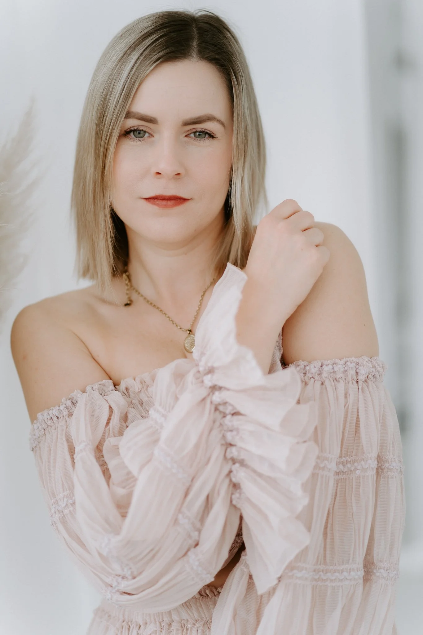 Eine junge Frau mit blonden, schulterlangen Haaren, die ein rosa, lockeres, ärmelloses Kleid trägt, steht vor einem weißen Hintergrund.