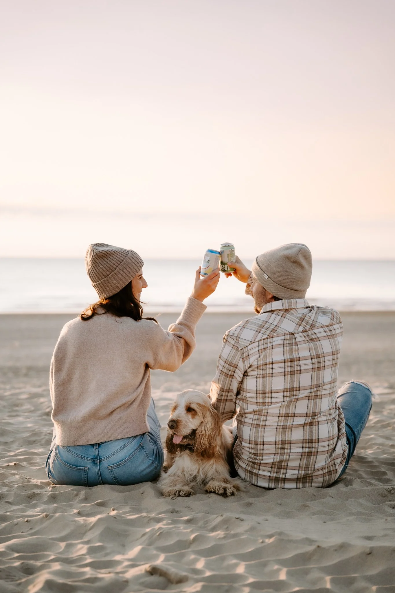 Ein Paar mit einem Hund sitzt am Strand und stößt beim Sonnenuntergang mit Dosen an.